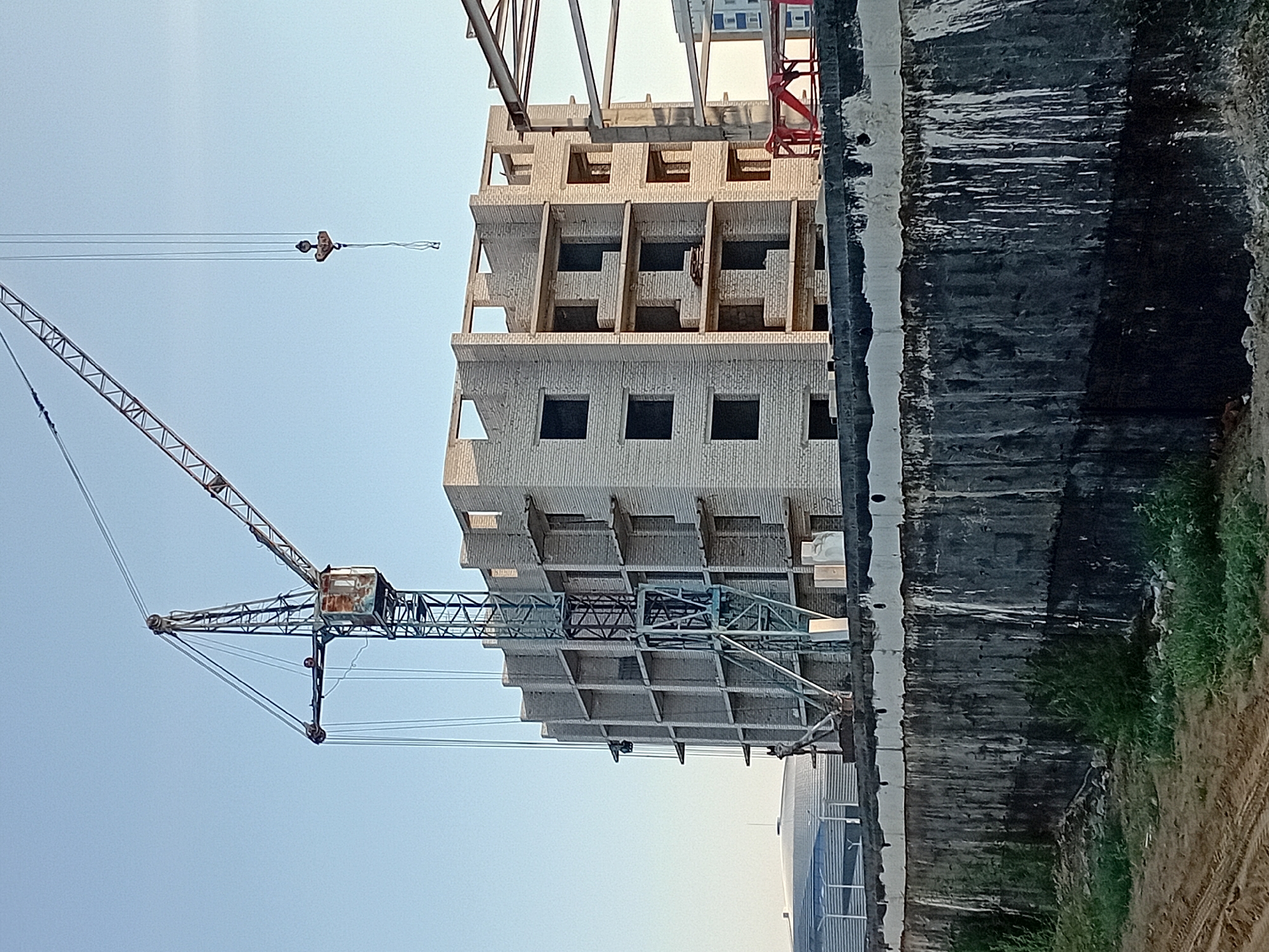 A construction site in the city of Semey, Kazakhstan, is depicted under a clear blue sky. In the foreground, a weathered concrete wall runs horizontally across the frame, exhibiting signs of age and wear with streaks of dark discoloration. Beyond the wall, a multi-story building is under construction, its light-colored brick walls forming a skeletal structure with visible openings for windows and balconies. A large tower crane, a prominent feature of the site, stands to the left of the building, its metal framework a stark contrast against the sky. The crane's cabin, weathered and aged, is visible at the top of its central mast. Another crane hook hangs from cables to the right, suspended in the air. In the background, a futuristic-looking domed structure with ribbed detailing is partially visible on the left. The ground in front of the concrete wall is a mix of sandy soil with tire tracks and patches of green vegetation.
