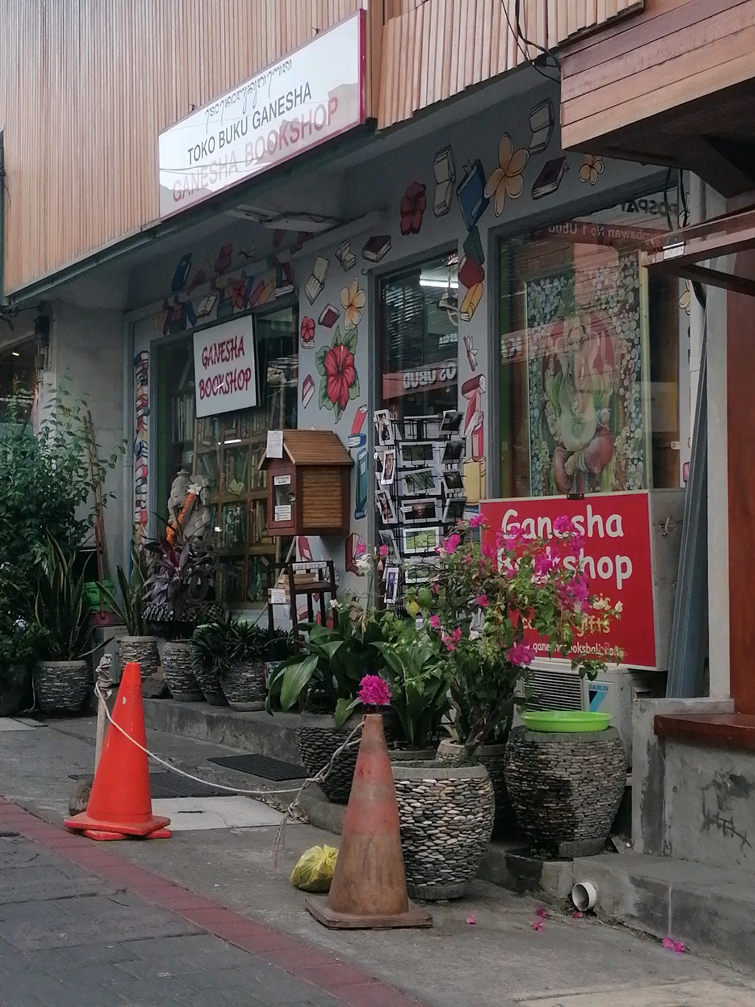 This image depicts the exterior of a bookshop named "Ganesha Bookshop" in Ubud District, Indonesia. The shop front features a prominent red sign with white lettering that reads "Ganesha Bookshop". Above this, a white sign with purple lettering says "Toko Buku Ganesha" and "GANESHA BOOKSHOP". The shop's facade is decorated with cartoonish drawings of books and flowers. Large glass windows reveal shelves stocked with books inside. In front of the shop, several potted plants are arranged, adding greenery to the scene. Two orange traffic cones are placed on the paved walkway, with one in the foreground and another slightly further back, both connected by a thin rope. A yellow plastic bag is visible near the second cone. The overall environment suggests a commercial street in Ubud, with traditional Balinese architectural elements visible in the building's facade and the use of natural materials like wood and stone in the planters.