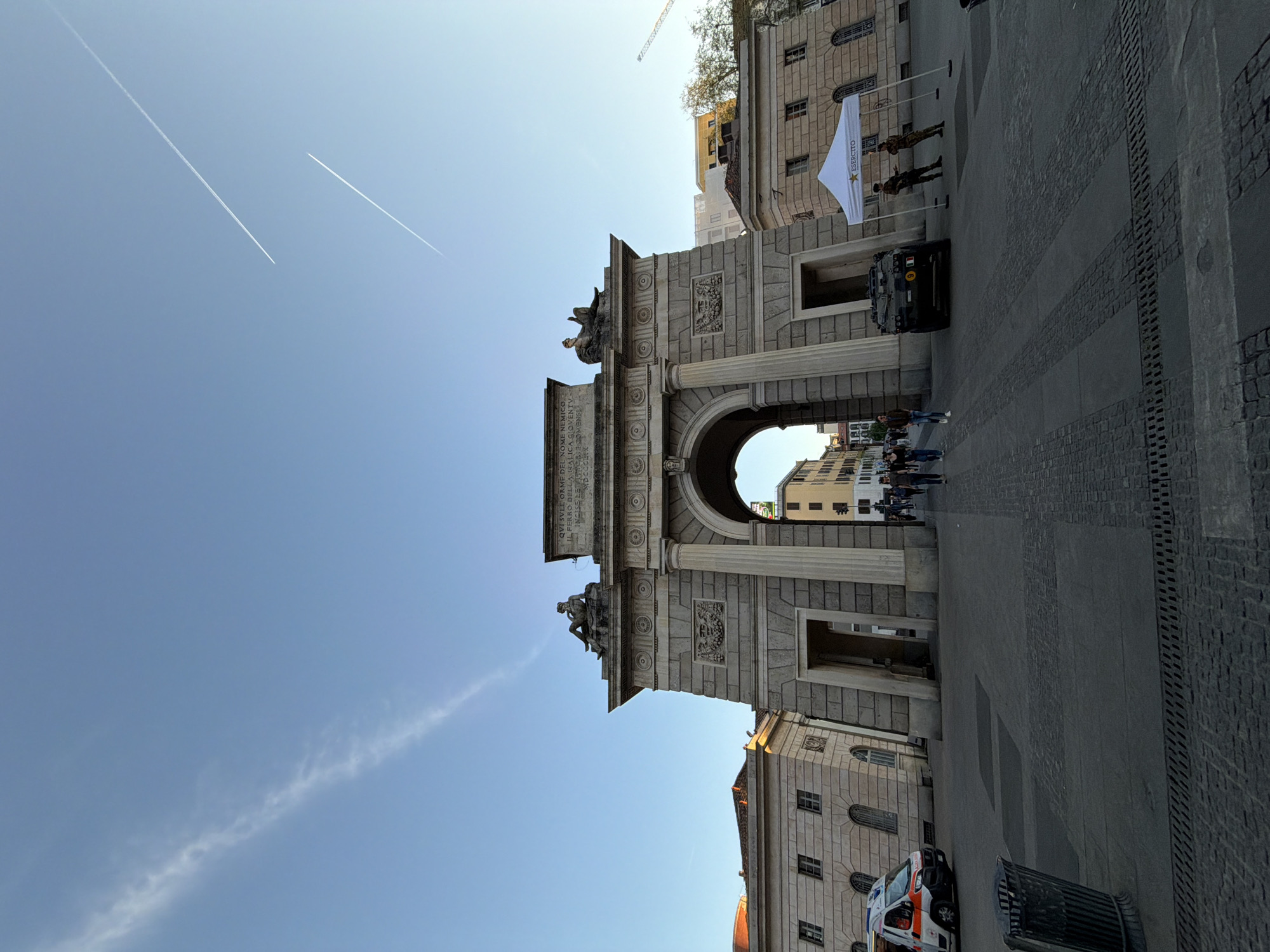 The image shows the Porta Sempione arch in Milan, Italy. The arch is made of stone and has statues on top. Two contrails in the sky, a building to the right of the arch. A military vehicle parked under the arch, soldiers standing near it, and pedestrians passing through. An ambulance parked on the right. A white tent is also visible near the vehicle.