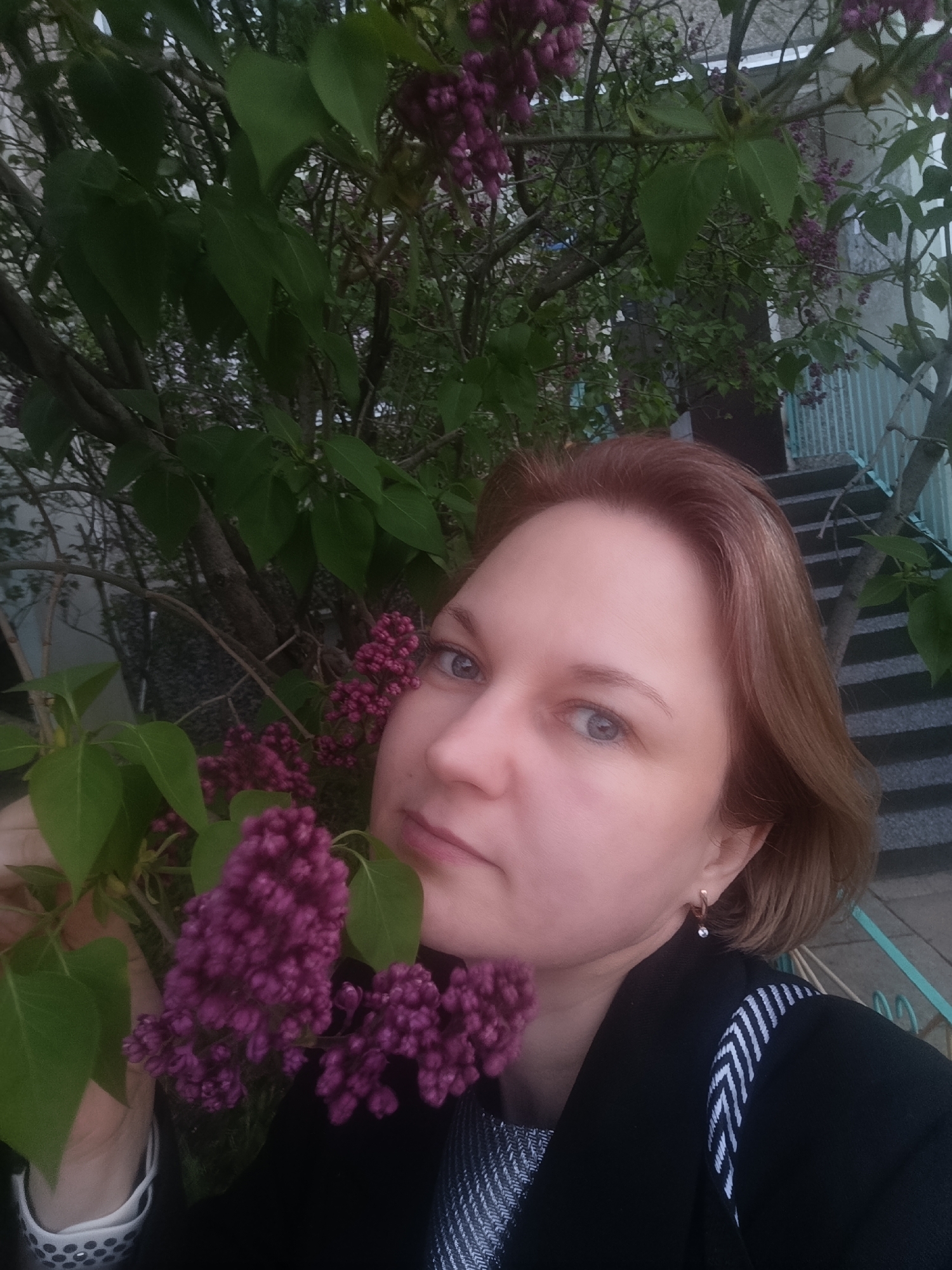A selfie taken in the city of Chaikovsky, Russia, features a fair-skinned woman with light brown hair styled in a bob, posing next to a lilac bush in bloom. She is wearing a dark jacket over a patterned top, and a smartwatch is visible on her wrist. The background includes a staircase with light blue railings and a light-colored building. The focus of the image is on the woman and the purple lilac flowers.