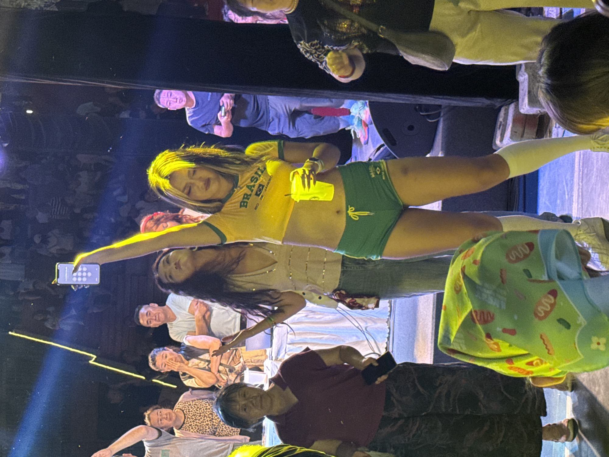 In Chaoyang District, China, at a stage event with a dark, crowded background, a young woman with blonde-streaked hair, wearing a yellow crop top with "BRASIL" printed on it and green shorts, is holding up a smartphone with her right arm. She is also holding a yellow cup in her left hand. In front of her, a light blue stage floor is visible, with some audience members in the foreground. The woman is illuminated by a bright yellow spotlight. To her left, a woman in a patterned top and dark pants is looking towards the stage. To her right, a man in a dark t-shirt and grey pants is visible. The scene appears to be a lively performance or event.