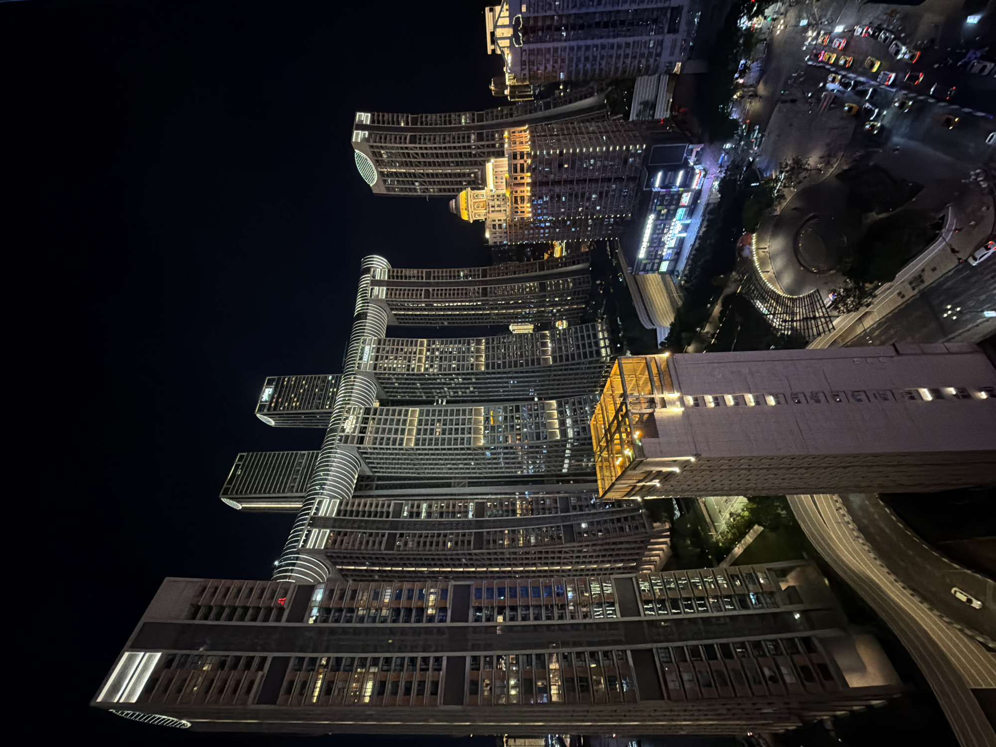 This image captures a nighttime view of the Raffles City Chongqing complex in Yuzhong District, China. The scene is dominated by the complex's distinctive architecture, featuring multiple skyscrapers connected by skybridges. The buildings are illuminated with lights, highlighting their modern design and creating a vibrant cityscape against the dark night sky. In the foreground, a multi-lane elevated road curves through the scene, with a white car visible on it. Further down, street-level traffic is visible, with numerous vehicles and illuminated storefronts. The overall impression is one of a bustling, contemporary urban environment.
