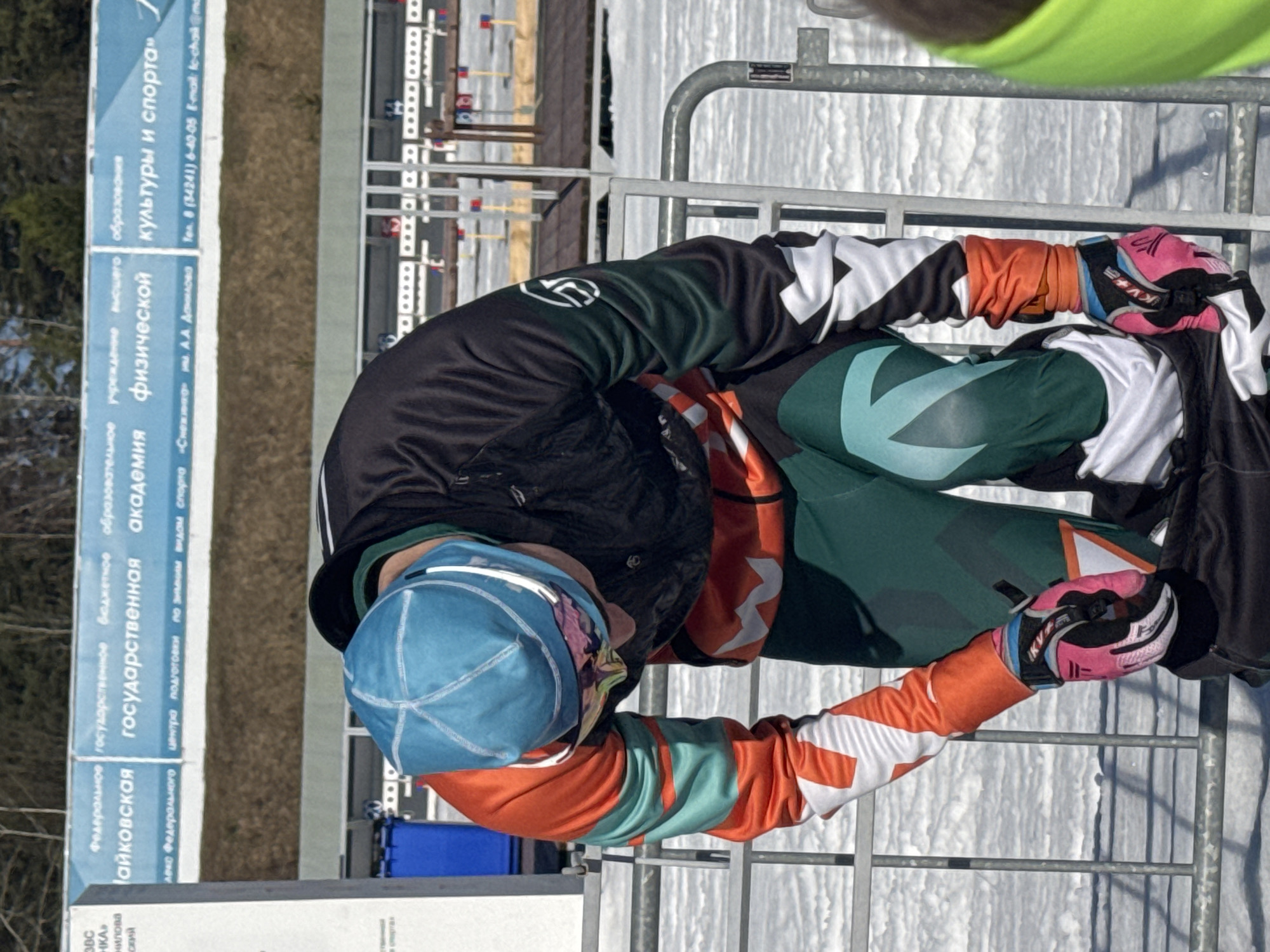 Here is a concise description of the image:
A person dressed in athletic wear (green and orange with white accents), wearing a light blue cap, is seen leaning on a metal barrier in what appears to be a winter sports facility. The background includes a biathlon shooting range. Signage in Russian is visible, identifying the location as the "Чайковская государственная академия физической культуры и спорта" (Tchaikovsky State Academy of Physical Culture and Sport) in Прикамский, Russia.