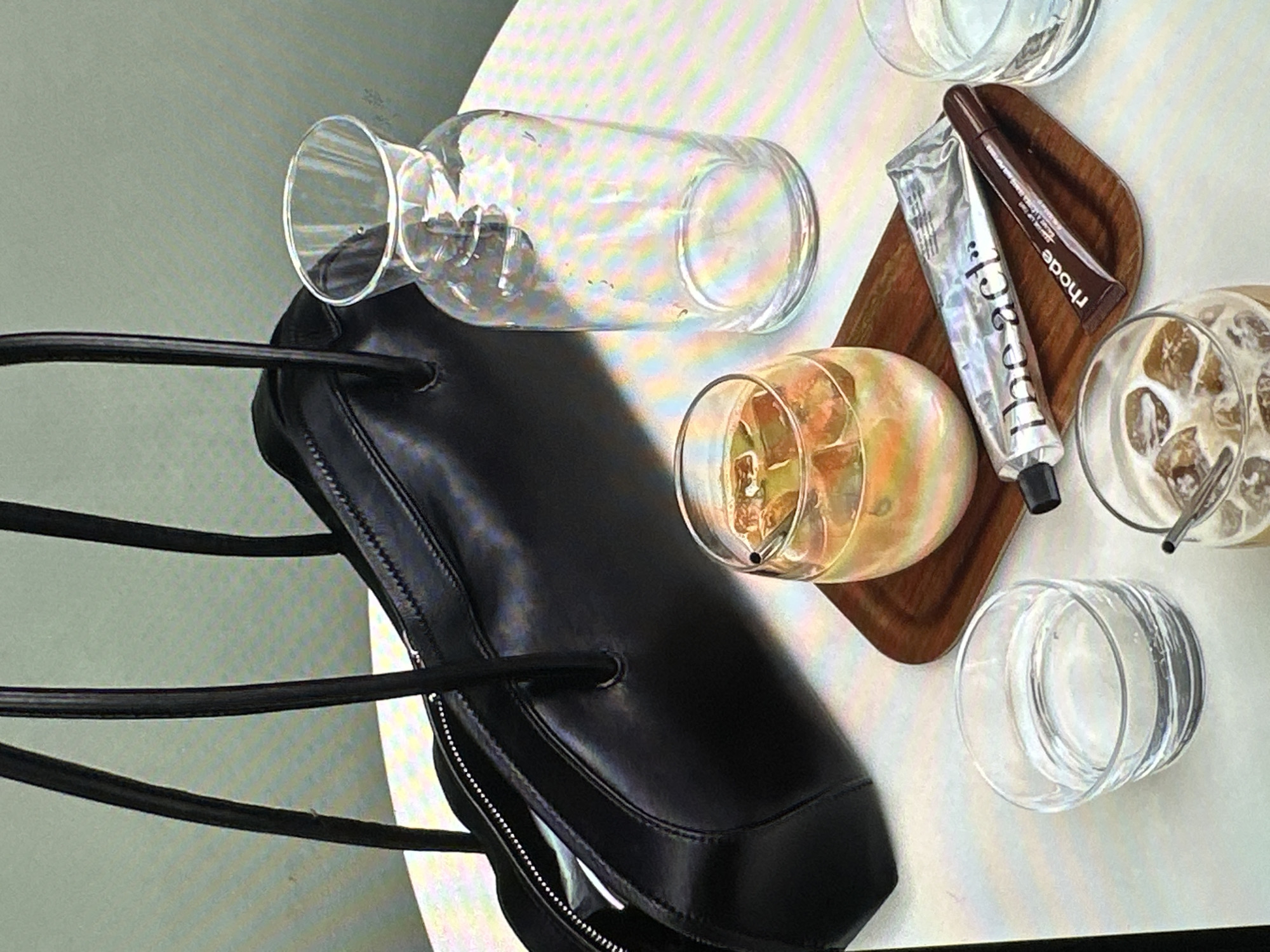 A high-angle shot captures a scene likely set in a cafe or indoor setting. A black leather handbag with a zipper closure and two straps rests on a surface. Beside the bag, there is a horizontal glass bottle and a glass containing what appears to be a cocktail with ice and fruit. A wooden tray holds a silver-colored tube of lip balm with black text and a brown-colored lip gloss with the text "rhode" on it, along with a glass of iced coffee with a straw. Another empty glass is also visible. The color palette consists of whites, creams, browns, blacks, and clear glass. The image is well-lit and does not suggest a specific location within Yerevan, Armenia.