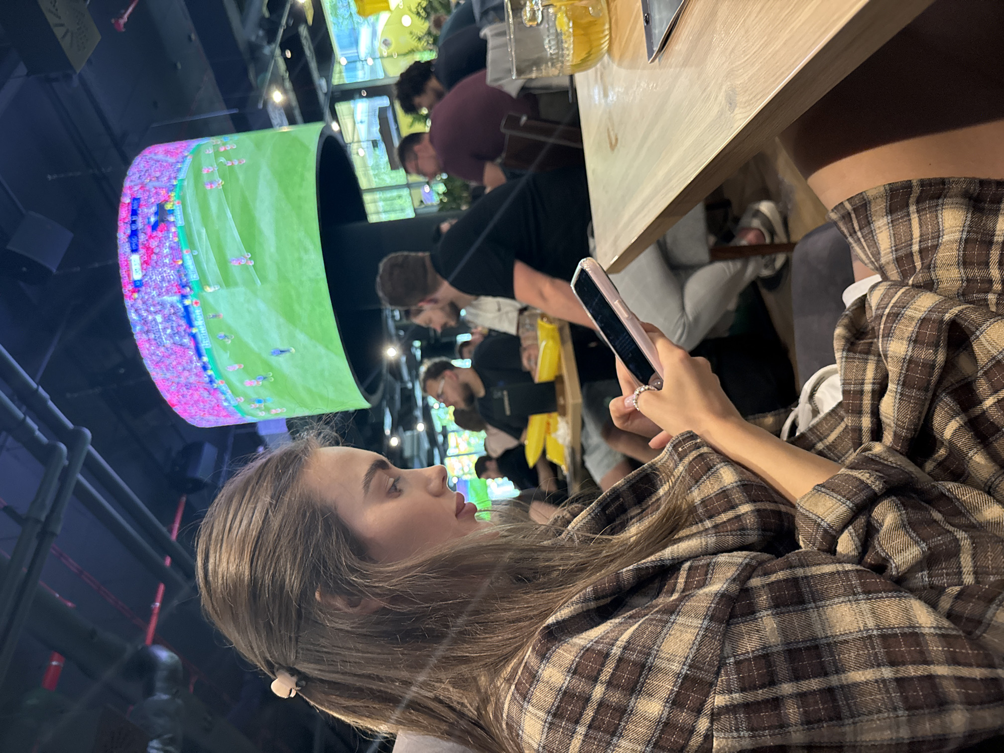 Here is a detailed description of the image:
The image captures a scene in what appears to be a bar or a public entertainment establishment with a large display screen. The main subject is a young woman sitting at a table, looking at her smartphone. She is wearing a brown and tan plaid shirt or jacket. Her hair is long and light brown, and she is facing towards the left side of the frame.
In the background, there are several people around a table, and a large, curved screen displays a brightly colored image that looks like a sports match, possibly football. Above the crowd, there are other screens visible, suggesting this is a place focused on broadcasting various events.
On the table, partially visible, there is a glass with a yellowish drink and possibly a menu or a tablet. The lighting is a mixture of ambient light from the screens and overhead fixtures, casting shadows and highlights across the scene.