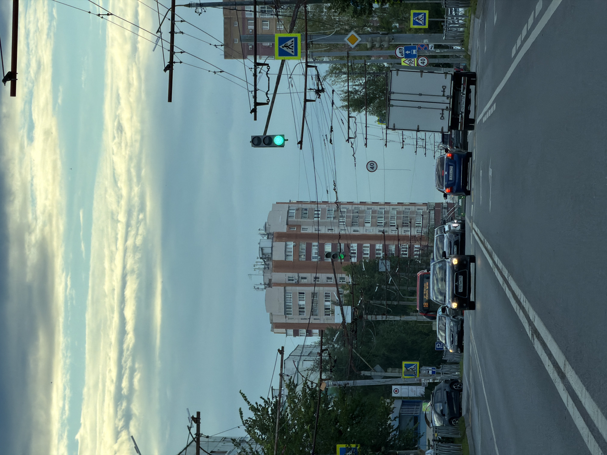 The image depicts a street scene in Kazan, Russia, likely during daylight hours with overcast skies. A multi-lane road stretches into the distance, with several vehicles in motion. The prominent feature on the right side of the road is a tall, modern apartment building with a facade of light-colored bricks and cream-colored sections. Adjacent to it, a traffic light shows a green signal. Overhead, a complex network of trolleybus wires crisscrosses the sky, supported by poles.
Several cars are visible on the road, including a black Mitsubishi Lancer in the foreground with its headlights on, a dark blue sedan, a white truck, and other vehicles further down the road. A red bus with the number "18" is also present. Traffic signs, including pedestrian crossings and speed limit signs (40 km/h), are visible. Green trees line the left side of the road, partially obscuring another apartment building. The sky is filled with layered clouds, some appearing lighter and some darker, suggesting a possibly humid or cloudy atmosphere. A few birds are perched on the overhead wires. The overall impression is of a busy urban environment.