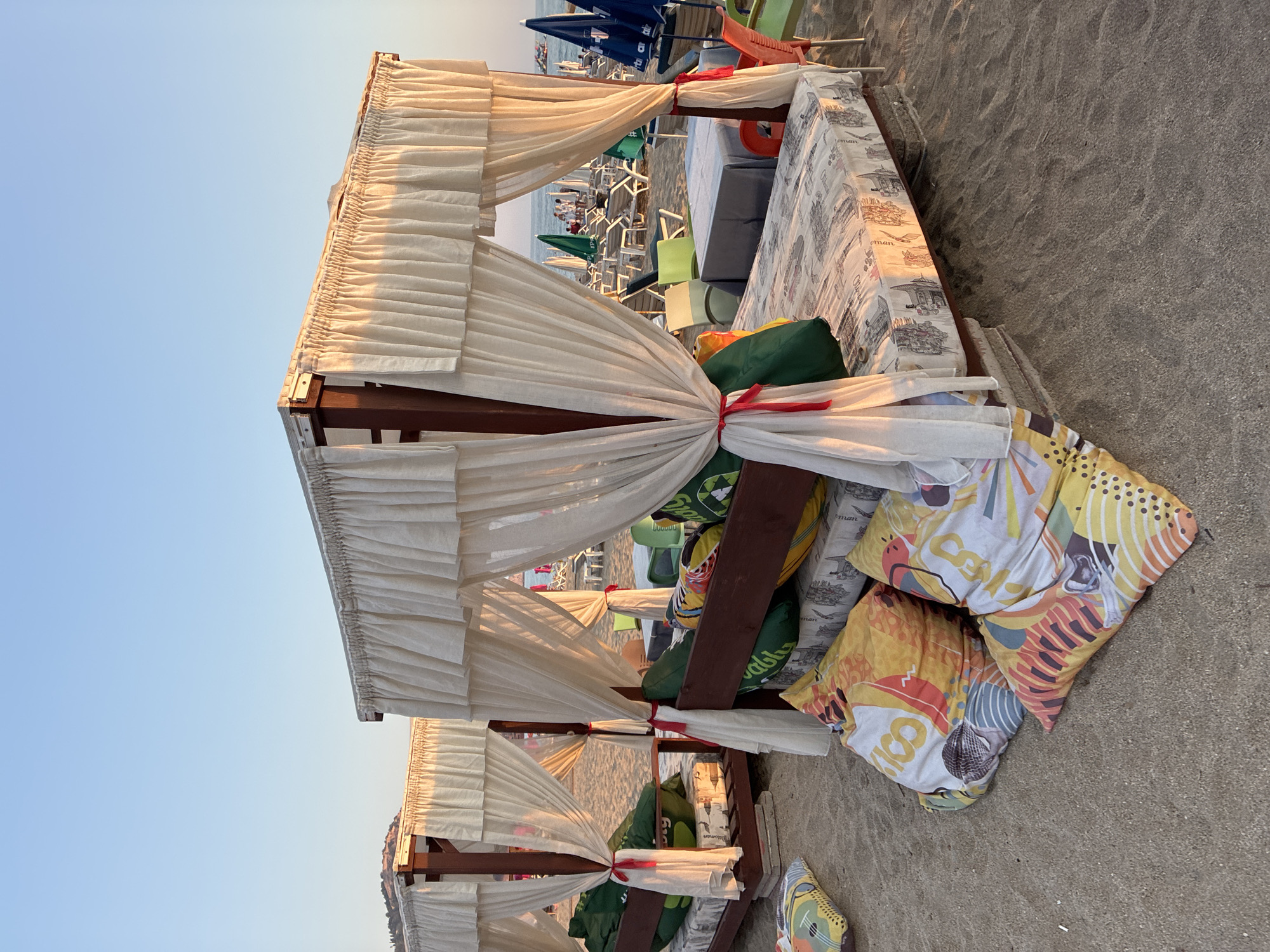 The image depicts a beach scene in Alanya, Turkey, during what appears to be late afternoon or early evening, given the warm, golden light. In the foreground, two elevated beach cabanas are prominently featured. These cabanas are constructed with dark wooden frames and are draped with flowing, cream-colored curtains tied back with red ribbons. Each cabana has a mattress on its platform, covered with a patterned textile. One cabana is closer to the viewer and shows colorful, large floor cushions scattered in front of it on the sand. The other cabana, slightly further back, is also furnished with green cushions.
Beyond the cabanas, the sandy beach extends towards the sea, which is visible in the background. The sea is calm, and several beach chairs and umbrellas are set up, some occupied by people. In the distance, a hill or mountainside is visible against the clear blue sky. The overall atmosphere suggests a relaxing and inviting beach resort setting.