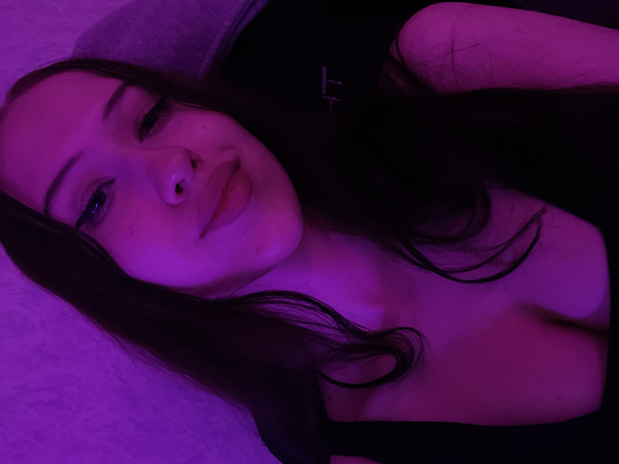 The media file is a close-up, overhead shot of a young woman lying down, illuminated by purple lighting. She has long, dark hair that cascades around her face and shoulders, and is wearing a dark-colored top. Her eyes are visible, and she appears to be looking towards the camera. The background consists of a textured surface, likely bedding, also tinted by the purple light. A small, faint symbol, possibly an embroidered logo, is visible on her top. The scene is intimate and dimly lit, suggesting a private indoor setting. There are no clear indications of time of day or weather conditions. The overall mood is relaxed and possibly moody, due to the lighting. There is no visible text other than the faint symbol on the clothing. The location context of Votkinsk, Russia, does not provide any specific visual cues within the image itself.