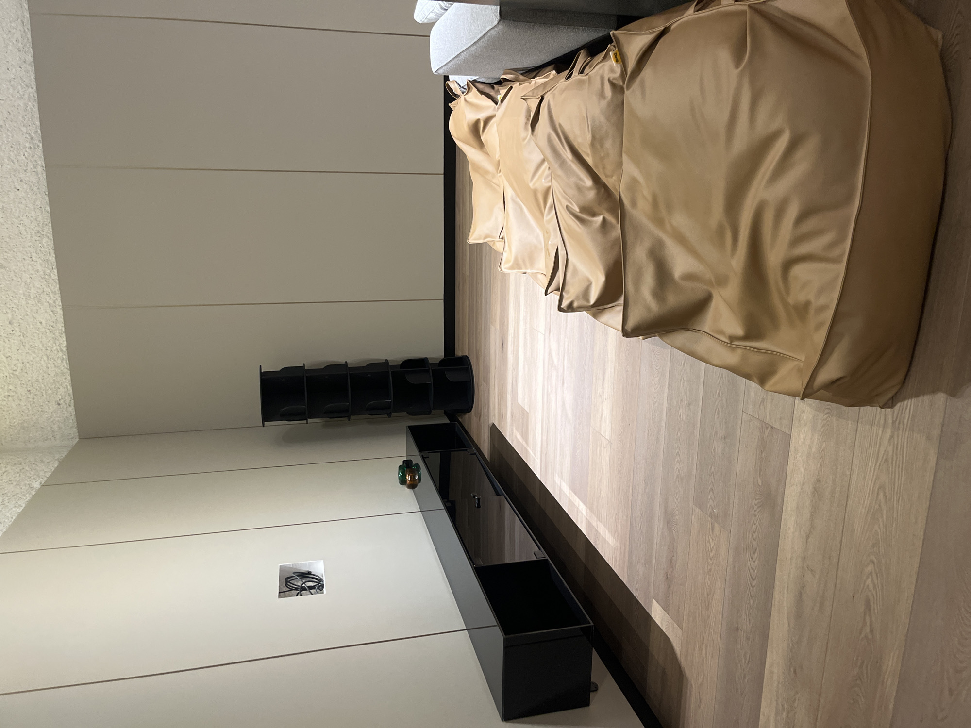 The image displays an interior room located in Mexico City, Mexico. The walls are covered in light beige paneling with vertical seams. The ceiling exhibits a textured, off-white finish. The floor is composed of light-toned wood planking. A glossy black, wall-mounted media console with two reflective cabinet doors is affixed to the left wall. Above the console, a square opening in the wall reveals coiled electrical wiring. A green and amber decorative glass object rests on the console's surface. A tall, black, cylindrical modular shelving unit is positioned against the back wall on the right. Six rectangular, tan faux leather bean bags, one marked with a "Pufy" tag, are arranged end-to-end on the floor, parallel to the wall, extending from the right towards the center. The end of a light grey upholstered sofa is partially visible on the far right. No people are present.
