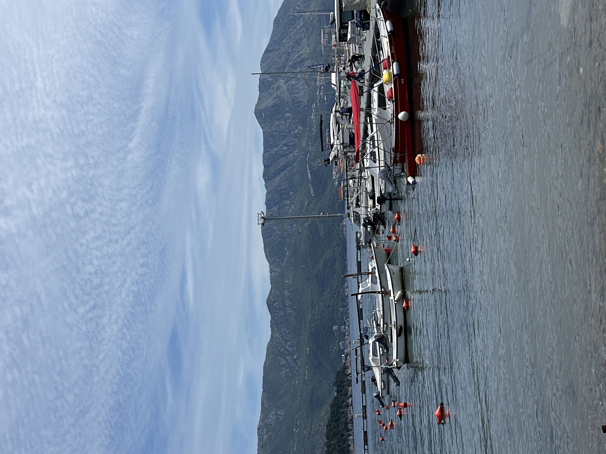 The image depicts a view from the city of Budva, Montenegro, showcasing a row of boats docked in the harbor. The boats vary in size and design, some with sails and others with motors. They are secured by orange buoys. Behind the boats, a mountainous landscape rises into the sky. The sky is partly cloudy. The water is relatively calm.