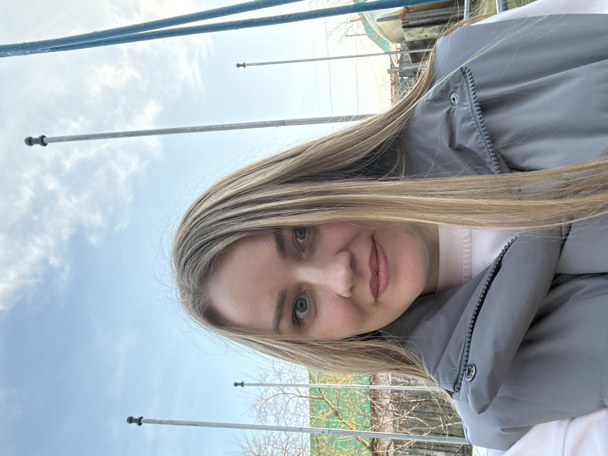 A selfie-style portrait captures a young woman, rotated 90 degrees counter-clockwise, making the horizon appear vertical. She has long, straight, light brown hair, blue-grey eyes, and a soft, pleasant smile. Her lips are lightly colored, and she wears minimal makeup. She is dressed in a grey padded jacket or vest with a visible zipper and a silver snap button, layered over a light-colored (possibly white or light pink) top.
The background reveals an outdoor setting under a bright, partly cloudy sky, indicating daytime. Several thin, metallic poles or rods are prominent, extending horizontally across the frame behind her, possibly part of a fence, railing, or other structure. In the upper right quadrant, there's a glimpse of what appears to be a light-colored boat or a section of an outdoor structure, and some bare tree branches suggest a late winter or early spring season. Further back, a section of green netting or tarp is visible. The overall scene suggests a casual moment captured in Chaikovsky, Russia, with elements that could be near a waterway or an outdoor recreational area. No visible text is present.