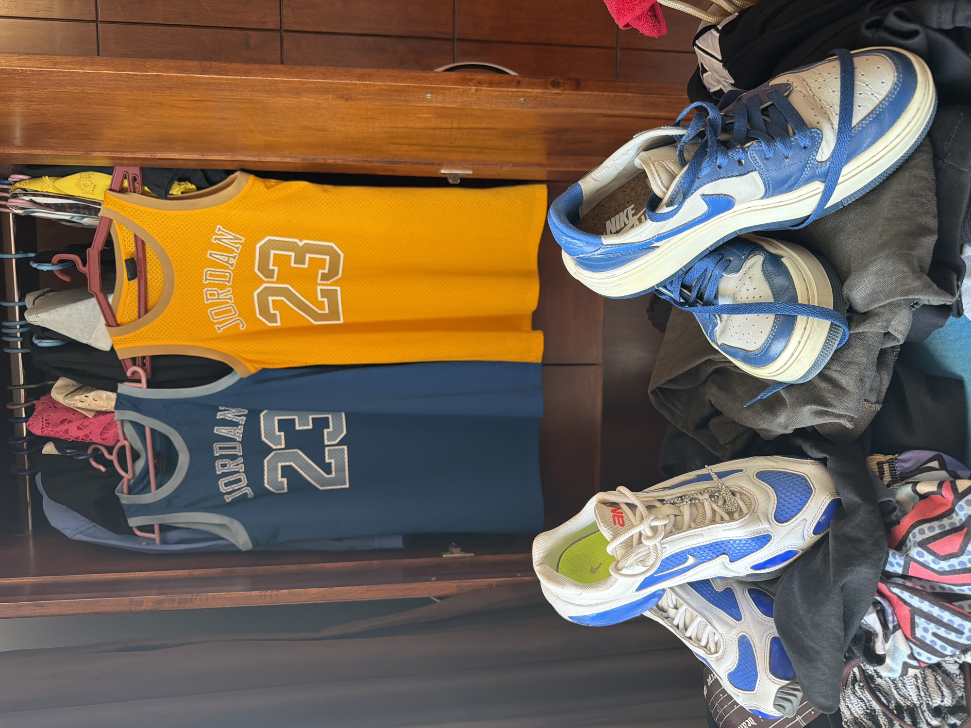 The image depicts the interior of a wooden wardrobe in Sharm El Sheikh, Egypt. On hangers, two basketball jerseys are displayed: one blue with "JORDAN 23" and one orange with "JORDAN 23". Below the jerseys, a pile of clothing is visible, with two pairs of athletic shoes placed on top. The left pair is white with blue accents, and the right pair is blue and white. The wardrobe appears to be filled with various garments.
