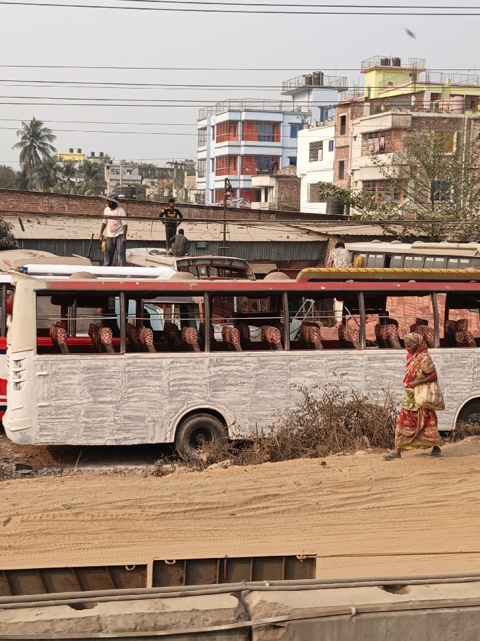 The scene in Boliarpur, Bangladesh, depicts an urban environment under a clear sky. In the foreground, a sandy embankment with visible tire tracks slopes downward, partially obscuring concrete barriers and metal structures at the bottom. A woman, dressed in a red and yellow sari with blue sandals, walks from left to right on this embankment, carrying a light-colored bag over her left shoulder.
In the midground, a white bus with a prominent red stripe is stationary on a dirt patch. Its exterior displays a textured, light grey finish, and rows of patterned brown seats are visible through its open windows. Dry shrubbery is situated to the bus's right. Multiple overhead utility lines crisscross the frame above the midground.
Behind the bus, several individuals are positioned on elevated structures. One man, wearing a light-colored shirt and a hat, stands upright on a roof-level structure, holding an indistinct object. Another man in a dark t-shirt and light pants stands further back on an adjacent roof. Two additional men are seated on different roof structures further to the right, one wearing a patterned shirt.
The background features several multi-story buildings of varying colors, including light blue, yellow, and white, some exhibiting balconies and rooftop water tanks. A single palm tree is visible on the far left background, amidst other sparse trees and vegetation.