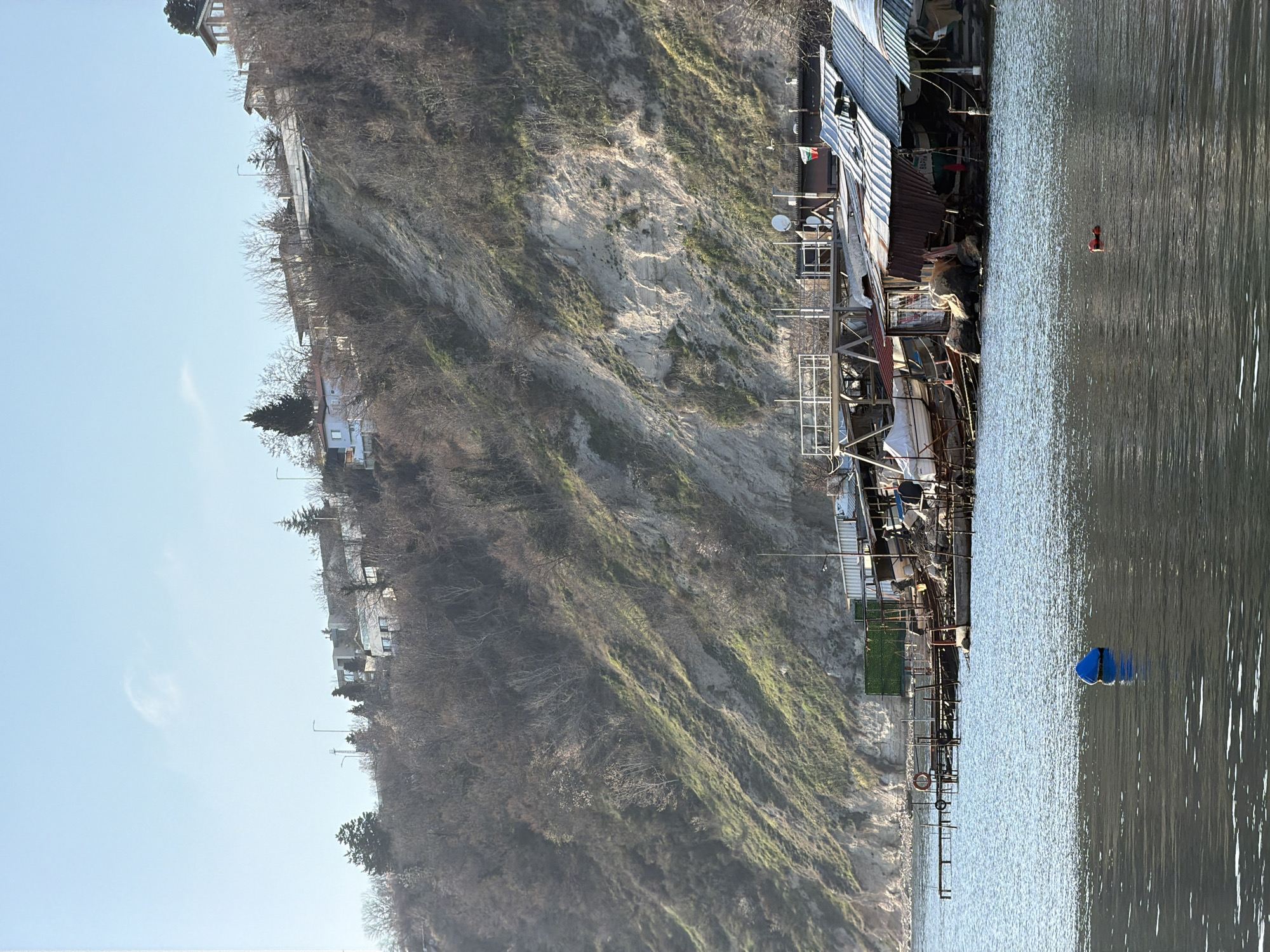 This image depicts a coastal scene in Varna, Bulgaria, characterized by water, shoreline structures, and a steep cliff. The foreground features choppy, dark blue water with a large, spherical blue buoy centrally located and a smaller, reddish-orange cylindrical buoy further right.
In the midground, a series of irregular, seemingly makeshift structures, possibly boathouses or fishing shacks, line the base of a steep cliff. These structures are built directly on the rocky shoreline and supported by wooden stilts and scaffolding. Their roofs are predominantly corrugated metal, with variations in color from rusty brown to weathered grey. Visible objects among these structures include several small boats, some covered with white tarps, and one white hull elevated on a wooden cradle. Multiple dilapidated wooden piers extend into the water, with one supporting a red and white life preserver ring. A green mesh screen is affixed to one of the buildings. A Bulgarian national flag (white, green, red horizontal stripes) flies from a pole on one roof, and multiple satellite dishes are mounted on various roofs. Text visible on one sign includes "BAPHA" (Varna in Cyrillic).
The background is dominated by a high, steep cliff face, exhibiting exposed light-colored earth and sand-colored sections interspersed with patches of sparse green vegetation. Numerous deciduous trees with bare branches, along with some evergreen trees, cover parts of the slope and the cliff crest. On the very top of the cliff, several residential buildings of varying architectural styles, some appearing modern with large windows, are visible. Streetlights are also present along the cliff's edge, suggesting a road. The sky above is clear and light blue with a few wispy clouds, indicating a bright, possibly cool, sunny day. No people are visible in the scene.