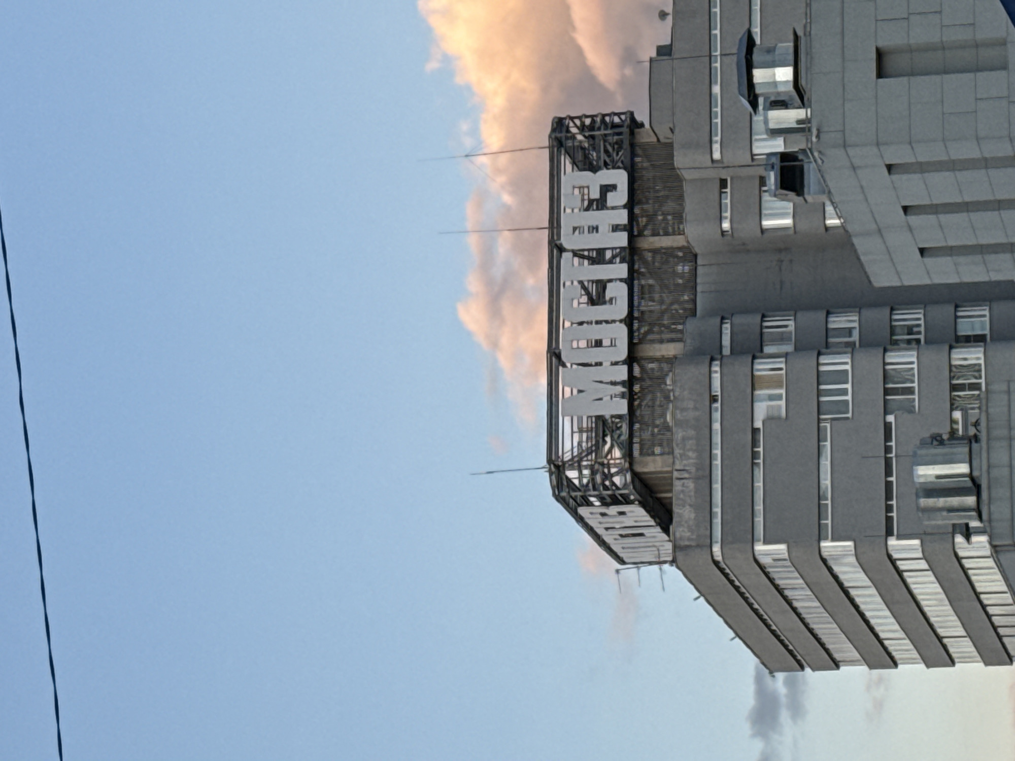 The image depicts the Izvestia building in Moscow, Russia, from a low-angle perspective. The building is a tall, concrete structure with a prominent "ИЗВЕСТИЯ" (Izvestia) sign on its upper portion. The sky is visible in the background, featuring a mix of light blue and soft, orange-tinged clouds. A thin, dark wire cuts across the leftmost portion of the frame, adding a minor element to the overall composition. The building's architecture features a distinct, stepped design, with multiple windows and balconies visible.