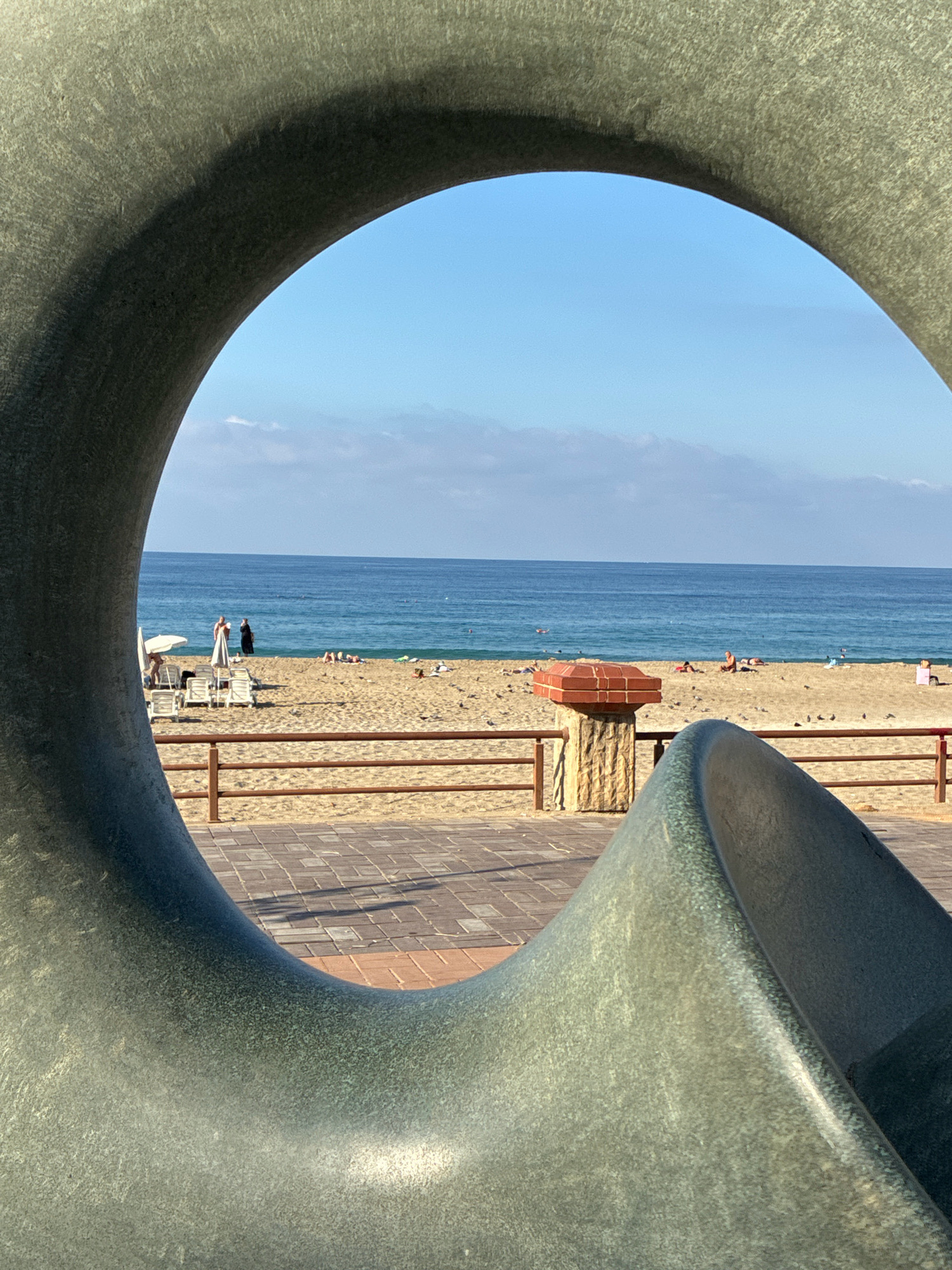 The image shows a view of a sandy beach and the sea framed by a large, abstract, gray sculpture. In the background, the calm, blue sea meets a hazy horizon. The beach is populated with several people, some lying on lounge chairs or towels, and others standing. A wooden fence separates the beach from a paved walkway. On the walkway, there are paving stones and shadows cast by an unseen object. To the left, white lounge chairs and umbrellas are set up on the sand. Further down the beach, a decorative concrete structure with a reddish-brown top is visible. The sky is clear blue with a few scattered white clouds. The sculpture in the foreground is textured and metallic-looking, with a curved, organic shape that obscures parts of the scene. The overall impression is a sunny day at a coastal location. The image is taken from a perspective looking through a large opening in the sculpture, framing the beach scene.