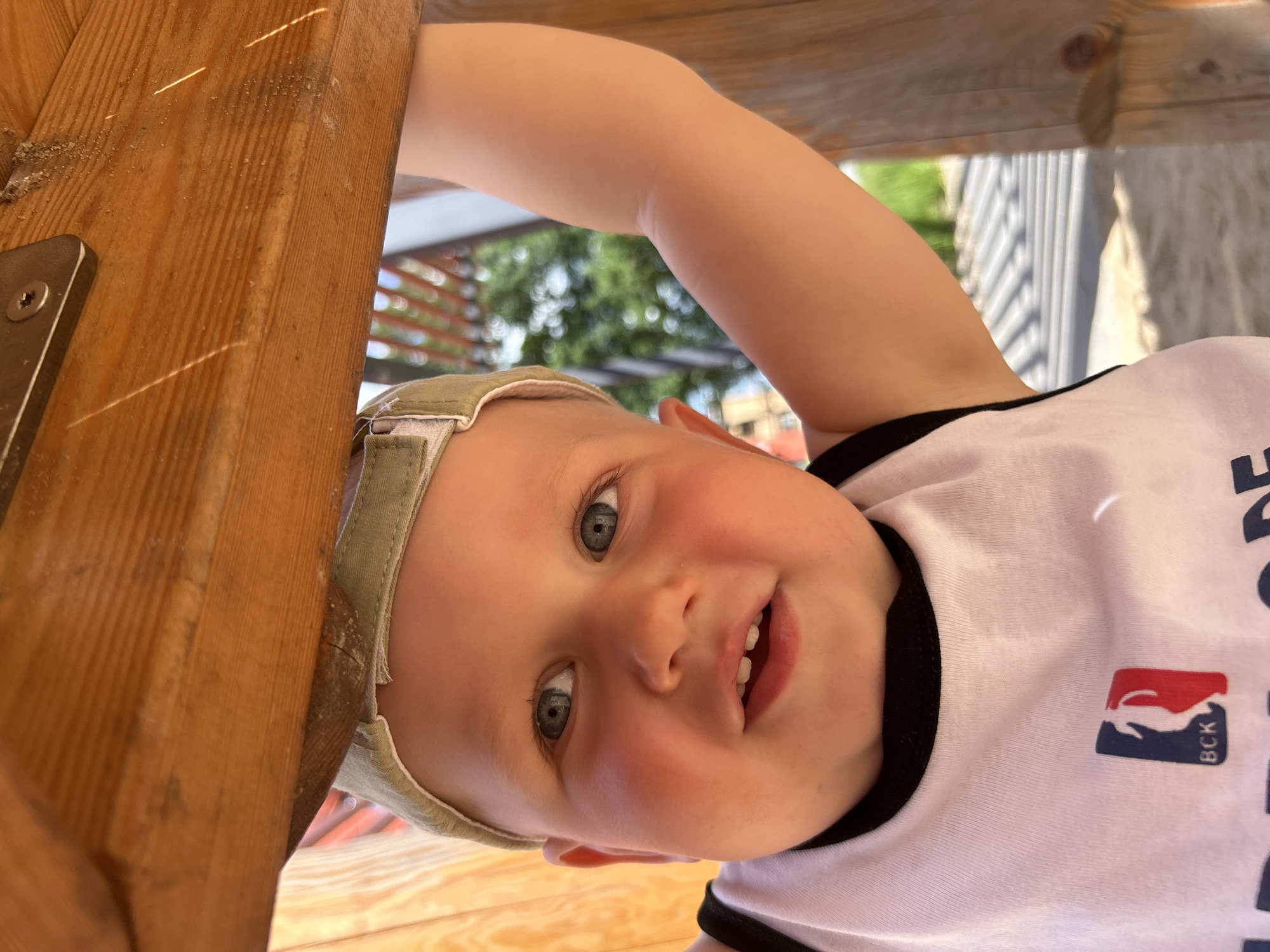 A close-up, eye-level shot features a young child, likely a toddler, wearing a light-colored t-shirt with a black trim and a cap. The child is looking directly at the camera with a slight smile. Their left arm is raised, gripping a wooden structure overhead, which appears to be part of a playground or outdoor seating area. The background shows blurred greenery and a striped wooden surface, suggesting an outdoor setting. The lighting indicates it is daytime, possibly sunny. There is no information about the location in Kaluga, Russia, within the image itself.