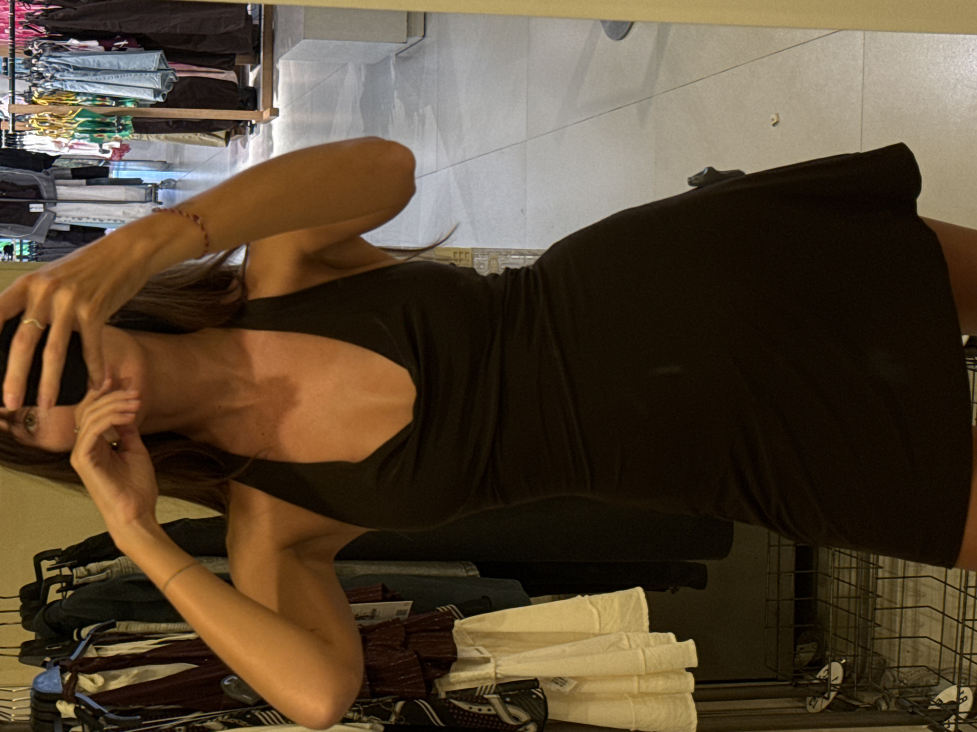 A woman is captured in what appears to be a mirror selfie, facing slightly to her left. She is wearing a dark, possibly black or dark brown, form-fitting dress with a V-neckline. Her right hand is raised to her face, covering her mouth and nose, revealing a gold-colored ring on her ring finger. Her left arm is visible, bent at the elbow, with a thin, multi-colored (red and white) bracelet on her wrist. Her long, dark hair falls over her shoulders.
The setting appears to be a retail environment or a large dressing area, likely in Kuta, Indonesia, as indicated by the location context. In the background, visible as a reflection, are several clothing racks laden with various garments, including jeans, colorful tops, and darker items. Below the racks, shelves hold stacks of folded clothes, some light-colored (possibly white or cream), and others patterned or darker. The floor is made of light-colored tiles. A metal grid structure, possibly a display basket or part of a fixture, is visible towards the lower right of the image. The overall lighting suggests an indoor environment during daytime or early evening, with artificial illumination. No discernible text is visible.
