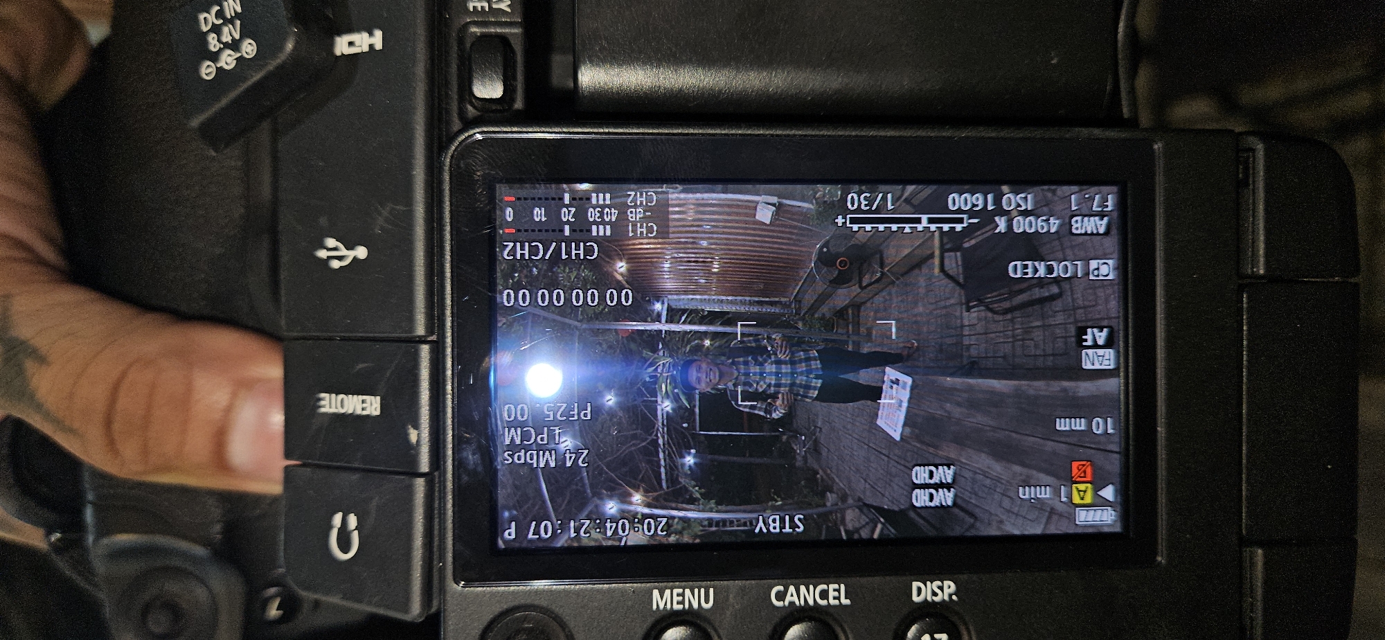 The media file displays a camera's rear screen, showing its interface and a live video feed, with parts of the camera body and a person's hand also visible.
In the live feed, a young man with a slight smile is the main subject. He wears a plaid shirt and dark trousers, standing on what appears to be a wooden deck or floor. The setting is outdoors at night or dusk, illuminated by numerous string lights, creating a festive ambiance. A corrugated metal roof is visible in the background, and there's a small fan attached to a structure.
The camera's display shows various settings and statuses:
* **Exposure:** Shutter speed 1/30, Aperture F7.1, ISO 1600.
* **White Balance:** AWB 4900 K.
* **Audio:** CH1 and CH2 levels are visible, indicated by bars.
* **Video Format:** PF25.00 (Progressive Frame at 25fps), LPCM (Linear Pulse Code Modulation audio), 24 Mbps (megabits per second), AVCHD.
* **Status:** The camera is in "STBY" (standby) mode.
* **Timecode:** "20:04:21:07 P" indicates an evening time (8:04 PM).
* **Lens:** Focal length is set to "10 mm".
* **Other indicators:** "AF" (Autofocus) is active, "CP LOCKED" (Control Panel Locked), a battery icon, and "1 min" (likely remaining recording time).
* **Buttons:** Below the screen, "MENU", "CANCEL", and "DISP" buttons are visible.
On the camera's body, various ports and their labels are discernible: "DC IN 8.4V", "HDMI", a USB symbol, and "REMOTE", along with a headphone symbol. The image provides a glimpse into a potential video recording setup in Bac Nha Trang Ward, Vietnam, during the evening.