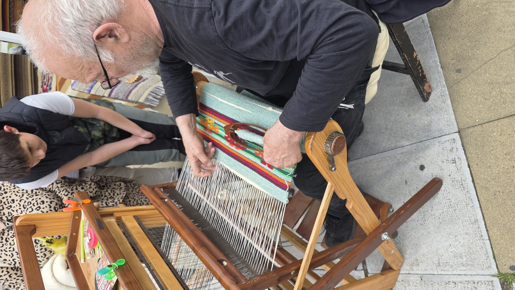 An elderly Caucasian man with white hair and glasses is weaving on a wooden loom. He is wearing a black long-sleeved shirt and black pants. His hands are positioned on the loom, working with the woven material. Beside him, a child wearing a black vest over a white shirt is seated, looking towards the man and the loom. The loom is set up on a sidewalk, adjacent to a paved area with visible grout lines. The woven piece features a pattern of turquoise with colorful horizontal stripes. The scene appears to be taking place outdoors during the day. There is no explicit information to confirm the location in Blagoevgrad, Bulgaria.