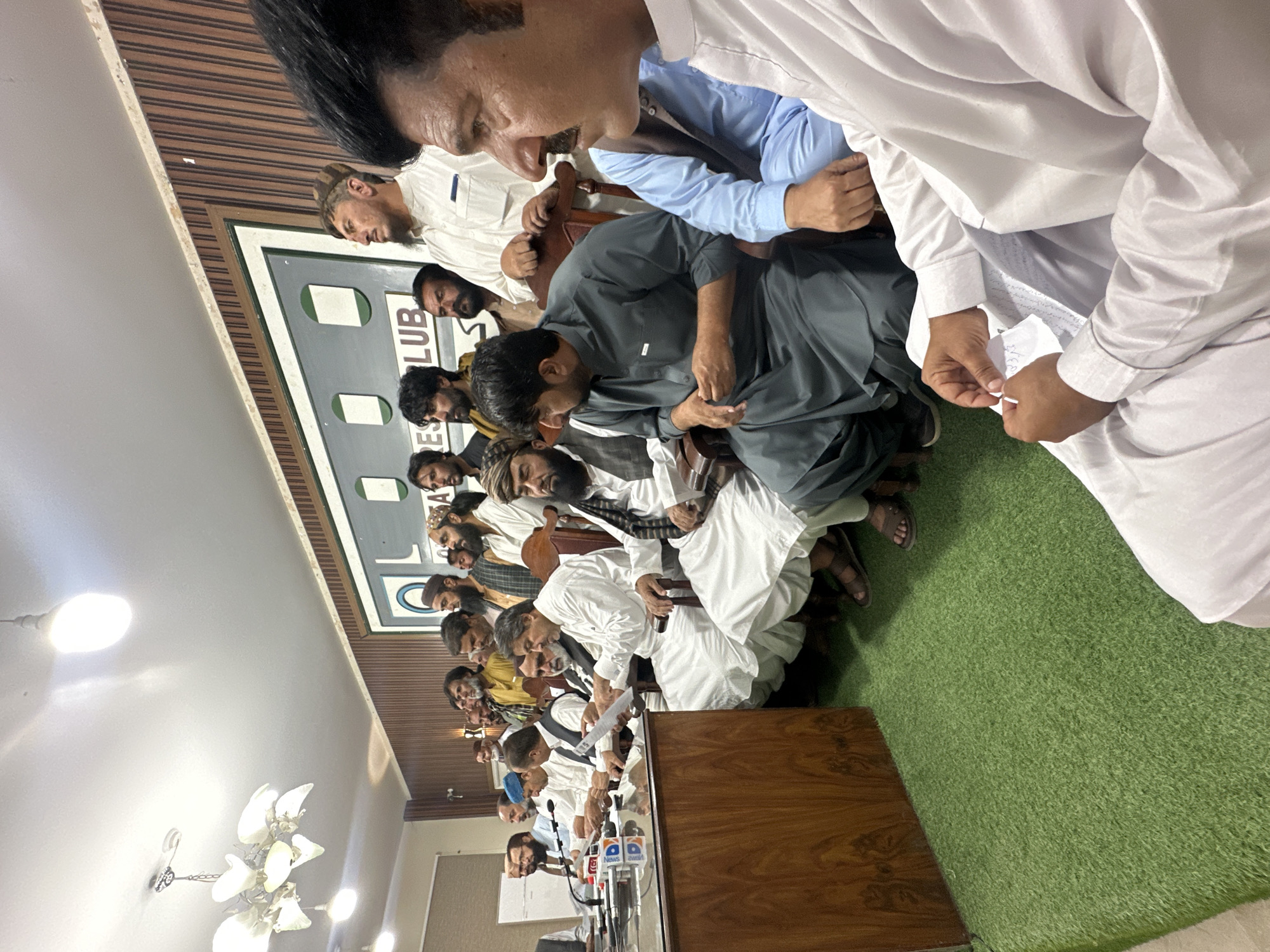The image shows a press conference in Quetta City Tehsil, Pakistan. A group of men, predominantly wearing traditional Pakistani attire (shalwar kameez), are seated at a long table. Microphones from various news outlets are visible in front of them. Some men stand or crouch behind the seated individuals. The room has a green carpet and wood-paneled walls. A sign with the word "CLUB" is partially visible.