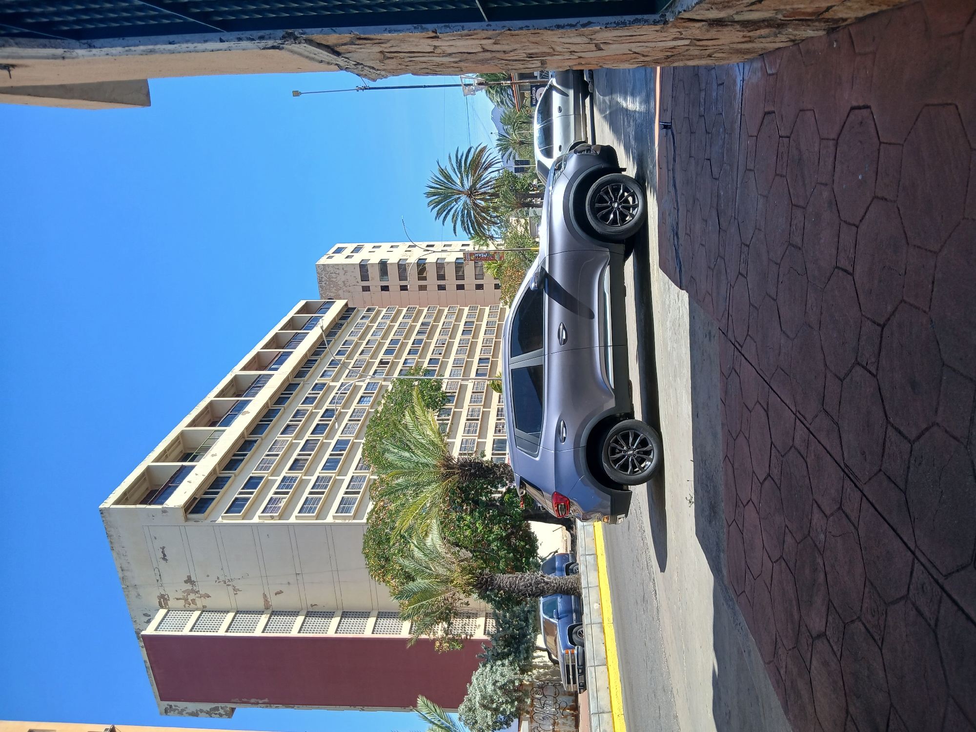 The image depicts a street scene in Juan Griego, Venezuela. A silver hatchback is parked on a street with a yellow line, partially obscuring other parked vehicles. In the background, there are tall, multi-story buildings with rows of windows. Palm trees are interspersed among the buildings. The sky is clear and blue. The foreground shows a textured, hexagonal-patterned sidewalk. The perspective is from within a partially enclosed structure, with visible sections of a ceiling and wall framing the view.