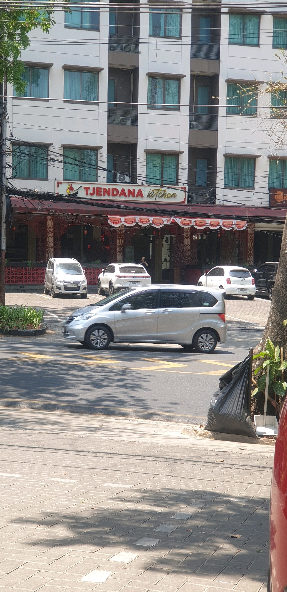 In Bandung City, Indonesia, a bright daytime scene unfolds with a multi-story building featuring white walls and numerous windows. A prominent sign with red lettering reads "TJENDANA kitchen." In front of the building, several cars are parked and driving. To the left, a white hatchback with license plate B 2076 QFL is parked. Next to it, a white SUV with license plate B 1511 YCV is also parked. A silver minivan is in motion, driving on the road. Further to the right, another white car with license plate B 1538 MCZ is parked, and a dark colored SUV is partially visible behind it. The street is paved with gray interlocking blocks. A black trash bag and a dustpan are placed on the sidewalk near a tree trunk. Lush green foliage is visible to the left of the building, and tree branches with sparse leaves are seen above. The sky is not visible in the frame.