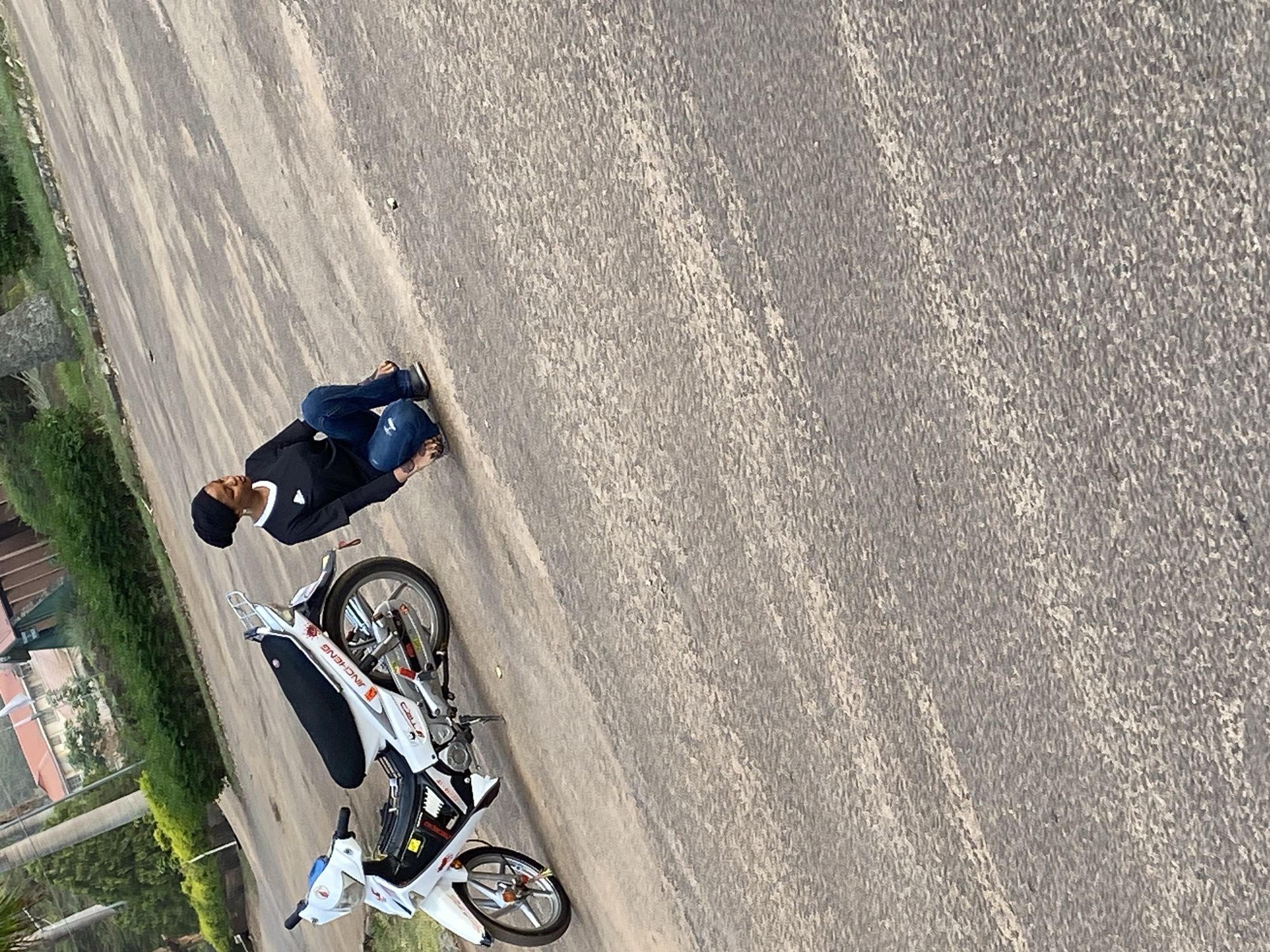 The image, rotated correctly for viewing, captures a scene on an asphalt road in Vom, Nigeria, during daylight. A single person is kneeling or crouching next to a white underbone motorcycle. The individual wears a black long-sleeved shirt with a white collar, dark blue jeans, a black head covering, and light-colored open-toed sandals.
The motorcycle is predominantly white with a black seat, silver spoked wheels, and various red and black accents. Visible text on the motorcycle includes "JINCHENG" printed in red below the seat, and "CELL" in white lettering on a red background near the rear wheel. Small red and gold decorative elements are attached near the front wheel's axle.
The setting appears to be outdoors on a textured grey asphalt surface, possibly a road or a large paved area. In the background, to the left, there is lush green vegetation consisting of bushes and trees. Partially visible through the greenery are light-colored buildings, suggesting a residential or institutional area. The lighting is bright, indicating a clear day with the sun high enough to cast distinct shadows.