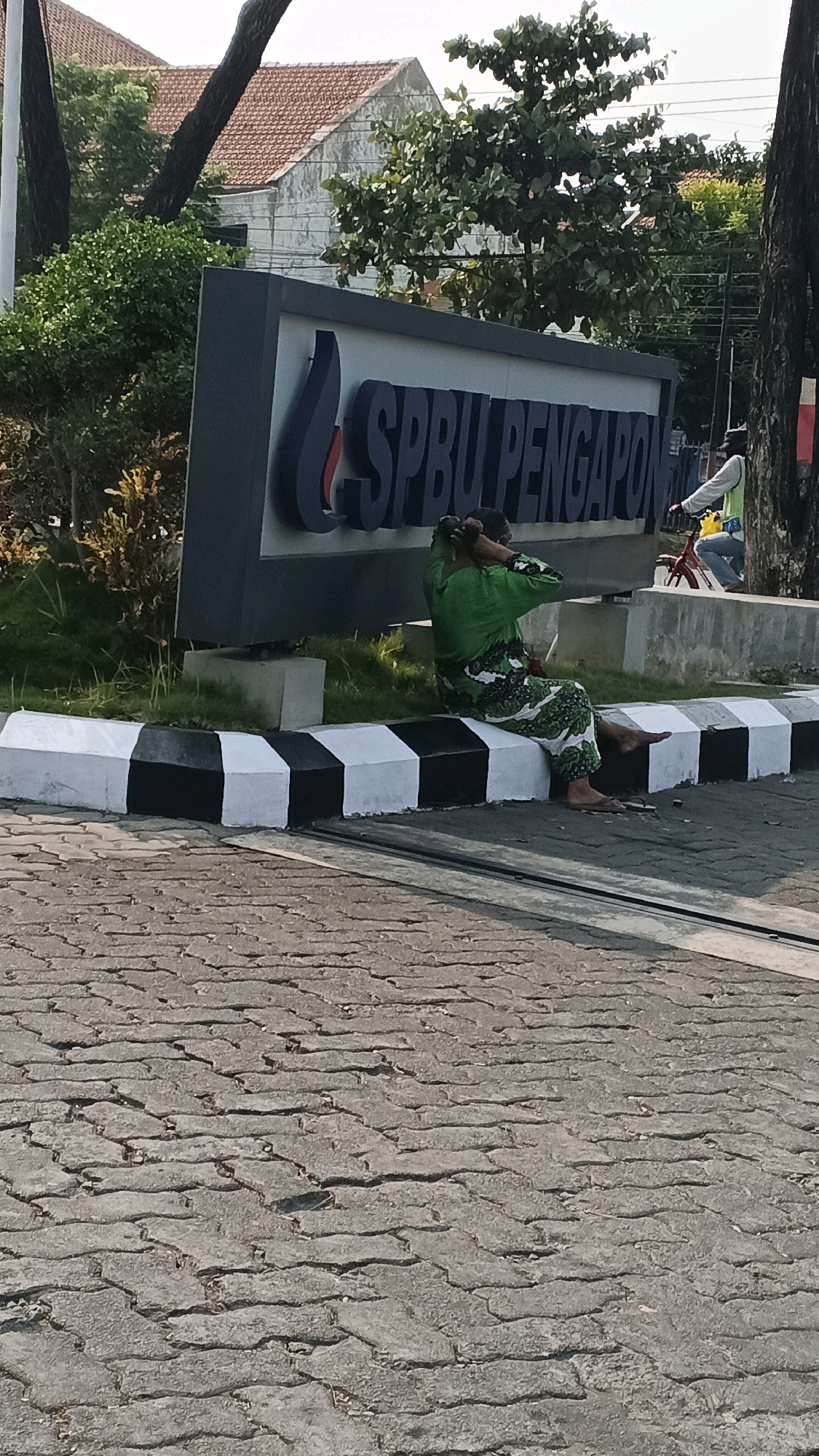 A woman is sitting on a black and white striped concrete barrier in front of a sign that reads "SPBU Pengapon". She is wearing a green and white patterned outfit and has her hands in her hair. The sign appears to be for a gas station and is located in the city of Semarang, Indonesia. Behind the sign, there are buildings and trees. The ground in front of the barrier is paved with interlocking concrete blocks. The lighting suggests it is daytime.