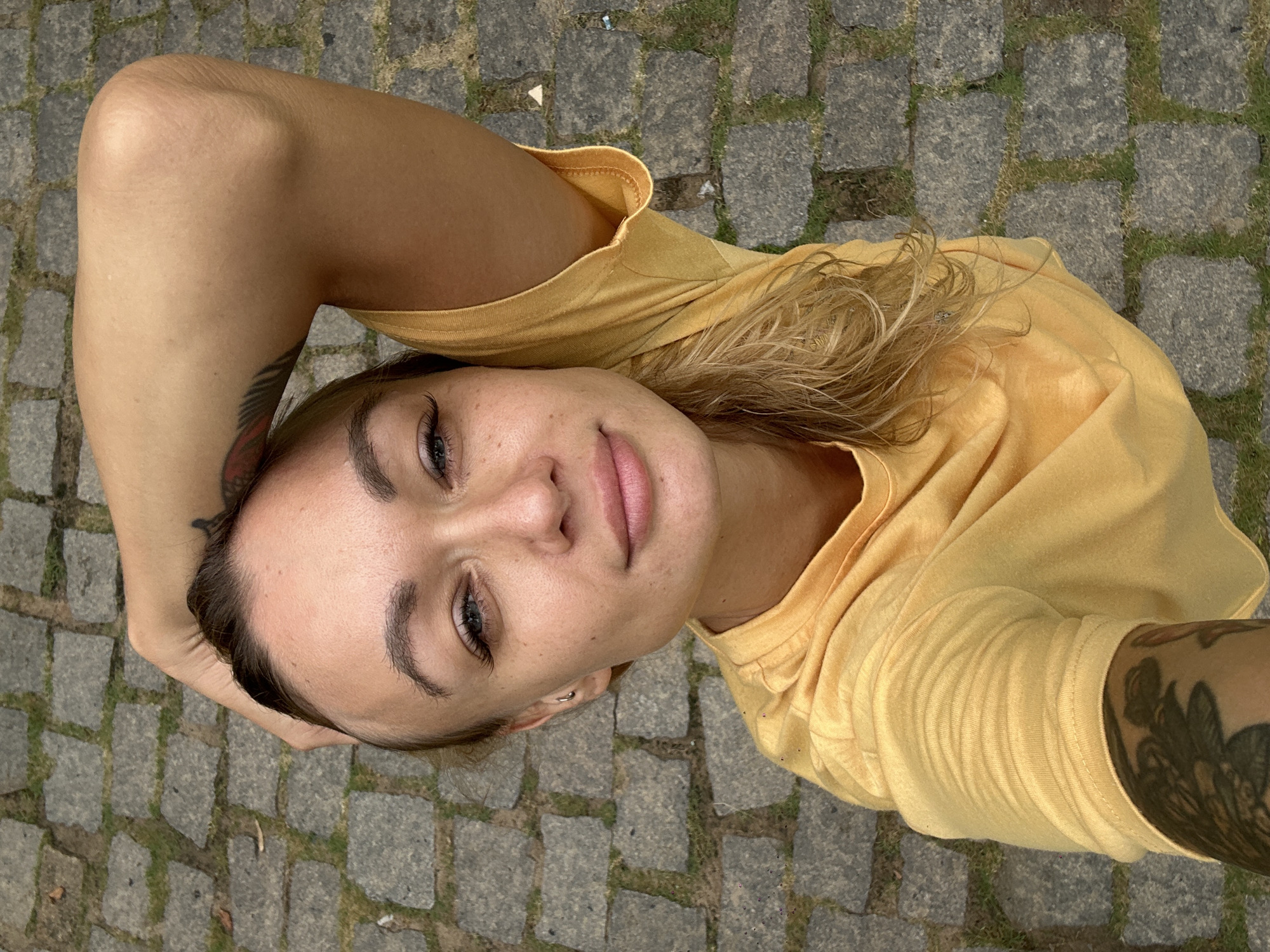 A woman in Phu Thuy Ward, Vietnam, lies on a cobblestone surface. Her yellow T-shirt is visible. The woman's face is turned upward, with her right arm raised above her head. Tattoos are visible on both arms.