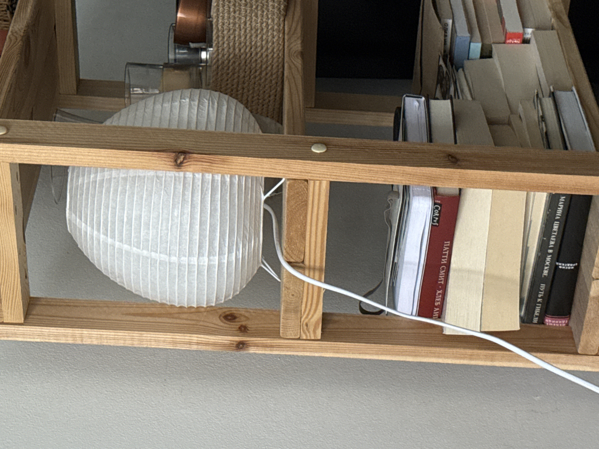 A white paper lantern is positioned on a wooden shelf. Next to it are two glasses, one containing a yellow liquid, and a copper bowl. Below the lantern, several books are stacked on lower shelves. The books have various colored covers, including red, cream, and black. The shelf unit is made of light-colored wood and stands against a gray wall. The image is a close-up shot, focusing on the shelf and its contents, with no people or animals visible. The location is not specified in the image.