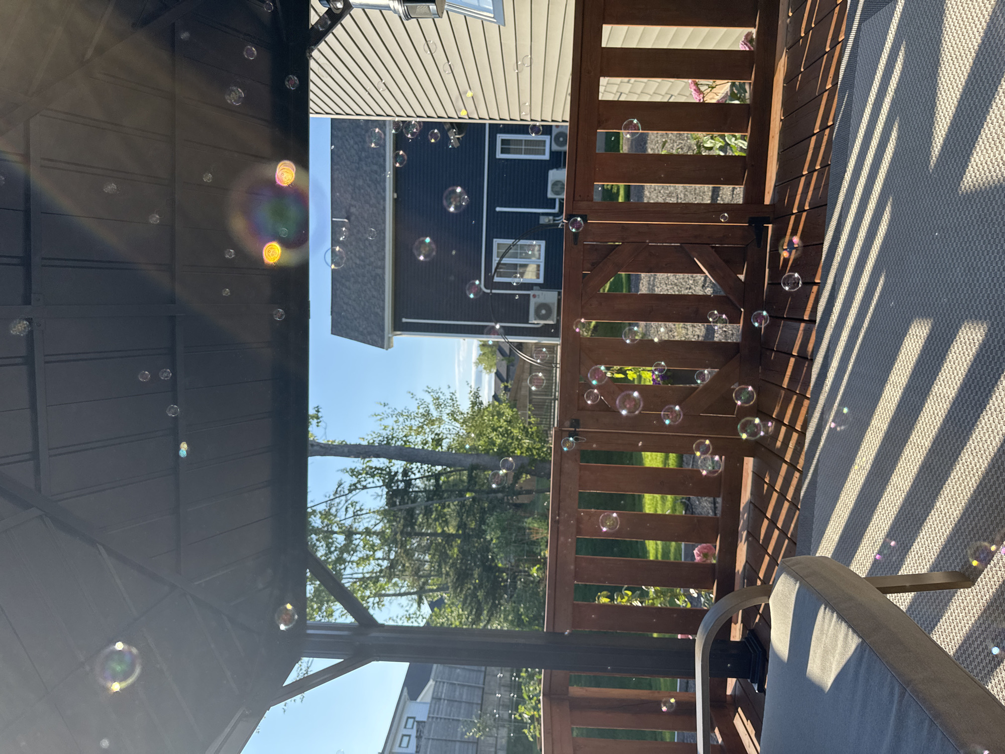 The image depicts a backyard deck with a gazebo. Numerous soap bubbles are floating in the air, some catching the sunlight and displaying iridescent colors. The deck is made of wooden planks and has a light-colored outdoor rug on one section. A gray outdoor chair is partially visible on the left side of the image. Beyond the wooden railing of the deck, a green lawn and trees are visible. Two houses are situated in the background, one dark blue and the other light-colored. The sky is clear and blue. The scene appears to be set in Dieppe, Canada, as indicated by the context of previous images. There are no people present in the image. The overall atmosphere is serene and suggests a warm, sunny day.