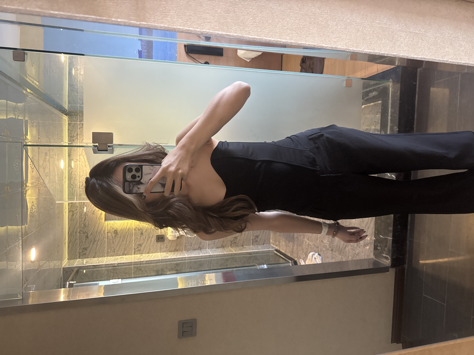 A person, likely female, is captured taking a mirror selfie in a modern bathroom. She stands, holding a smartphone with a light-colored case up to obscure her face. She wears a form-fitting black strapless top and black trousers. Her long, dark brown hair cascades over her shoulders and arm. A white or light-colored wristband and several beaded bracelets are visible on her right wrist.
The bathroom features light-colored, possibly marble-tiled walls, with recessed lighting fixtures reflecting in the mirror. Glass shelves or partitions are also visible. The flooring appears to be dark, polished stone or tile. In the background reflections, through what seems to be a window or opening, a blue sky or body of water is visible. Further inside, reflections show what might be a desk with a monitor and cables, suggesting the bathroom is part of a larger room, possibly in a hotel or a modern apartment.
The activity is a self-portrait taken using a mirror. The visible outdoor light suggests it is daytime. The location context provided is Ganzhou, China. The style of the bathroom and the implied larger setting are consistent with modern urban environments. Text on the phone case and wristband is present but illegible.
