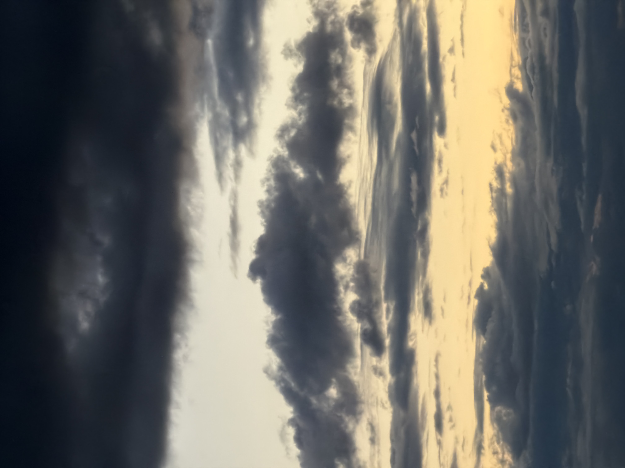 The image shows a sky filled with dark, heavy clouds illuminated by a yellow light source, possibly sunset or sunrise. The sky is predominantly a mix of gray and black clouds, with strips of yellow light interspersed, giving the impression of a stormy or dramatic weather scene. There are no people, subjects, or objects present in the frame. The location and city are unknown.