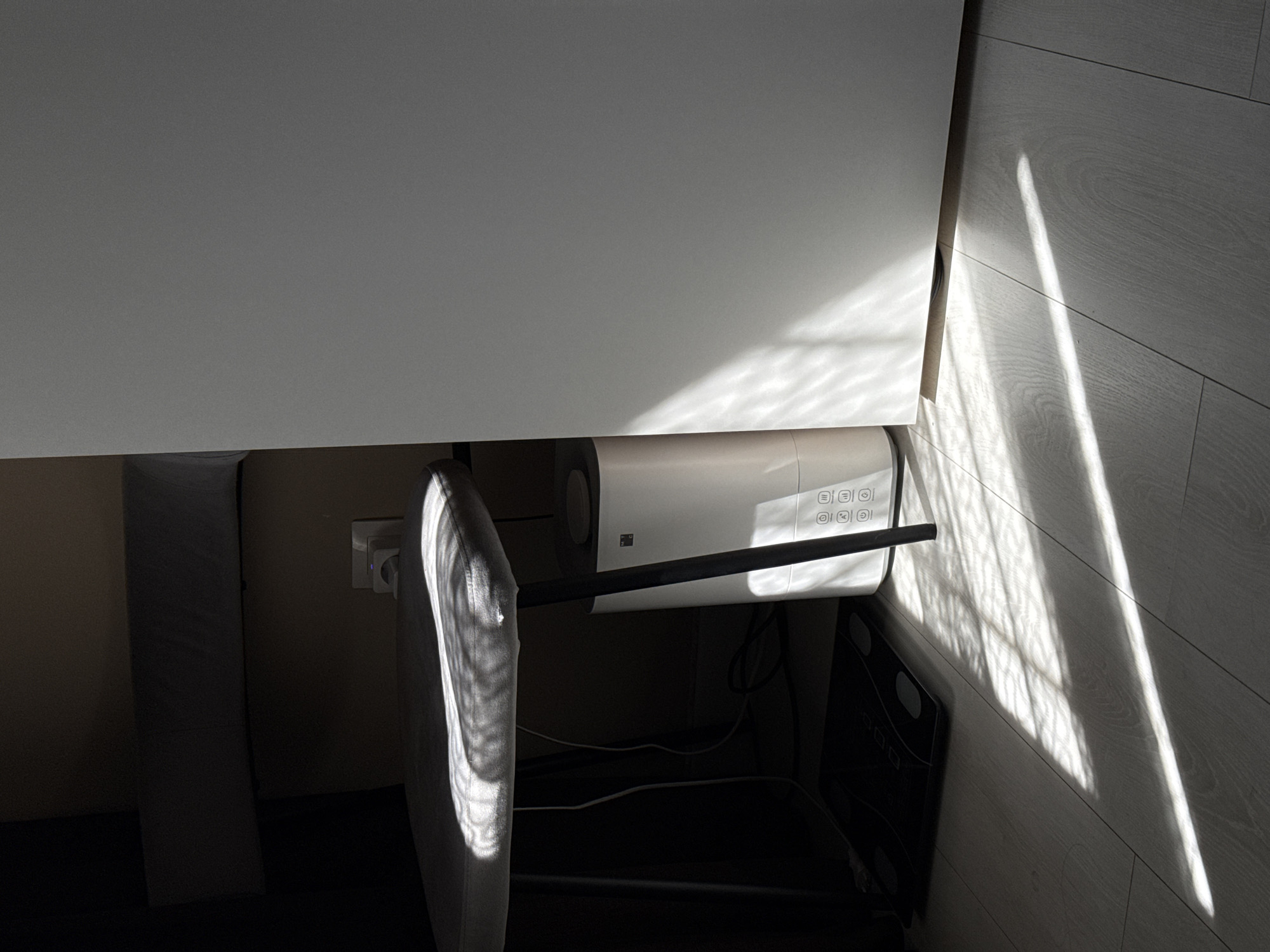 The image depicts an indoor scene, likely a home office or personal space, illuminated by strong natural light. A large, white desk or table occupies the upper part of the frame, casting a prominent shadow over the area beneath.
Underneath the desk, a white, rectangular electronic appliance is visible, possibly an air purifier or similar device. It features a control panel with a grid of circular buttons or indicators, displaying various symbols such as an "O" shape, an "A" within a circle, and what appear to be fan or mode icons. A distinct square symbol with four smaller squares inside is also present on its surface. A black horizontal bar runs across the front of this appliance, suggesting it is part of its stand or a structural element.
To the left, a grey, upholstered surface, likely part of a chair or stool, catches some light. Further left, a power outlet is visible on a light beige wall, with a plug inserted. The floor consists of light-colored wooden or laminate planks, exhibiting clear grain patterns. On the far right, a dark digital scale is partially visible on the floor. Various electrical wires are loosely spread across the floor underneath the desk.
The scene is characterized by dramatic lighting, with sharp, elongated diagonal shadows cast by the sunlight streaming in, likely from a window not visible in the frame. This strong interplay of light and shadow suggests either early morning or late afternoon on a clear, sunny day. There are no people visible, and no specific activity is taking place; the scene portrays a static interior setup.