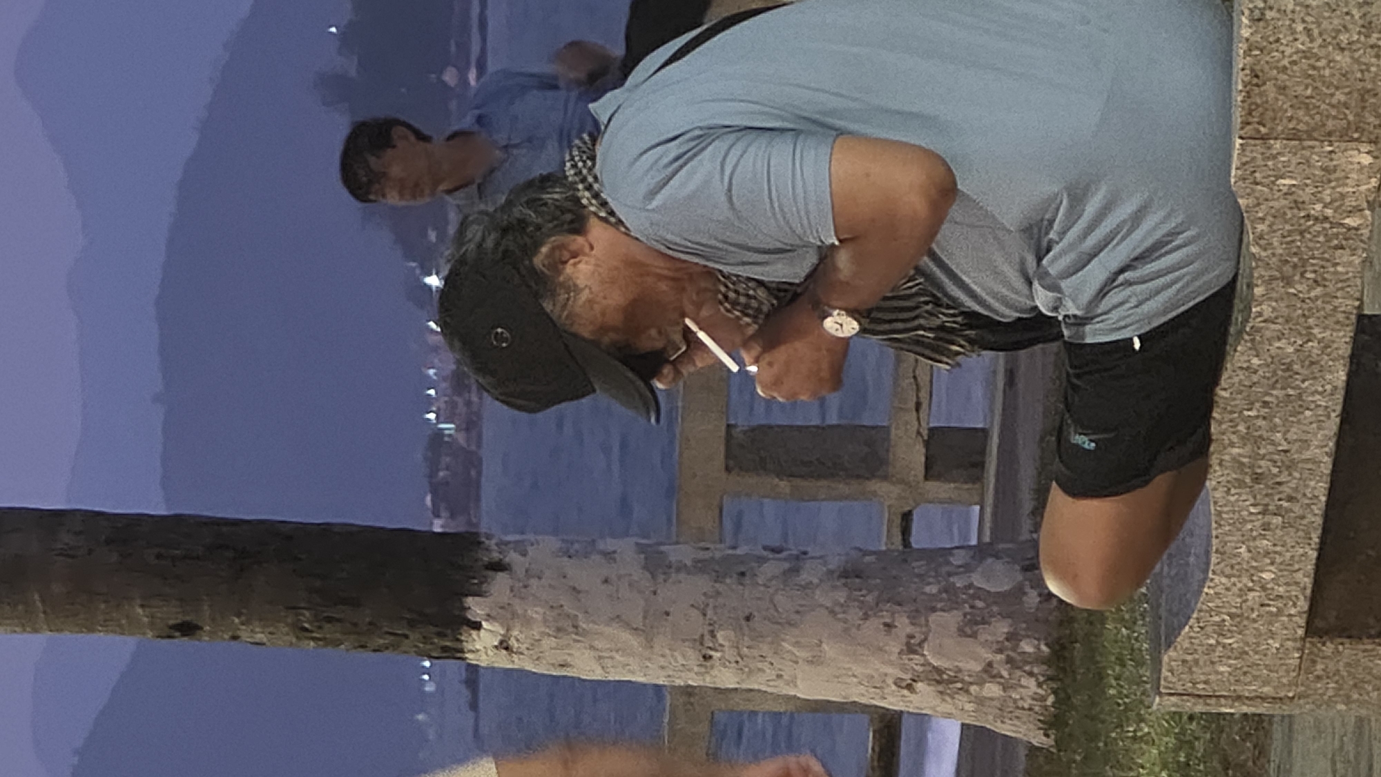 An adult male is smoking a cigarette near the shore in Nha Trang, Vietnam. He is wearing a light blue short-sleeve shirt, black shorts, a scarf around his neck, and a black baseball cap. He is leaning slightly forward, his head lowered as he smokes. A wristwatch is visible on his left wrist. In the background, there is a body of water, the city lights are visible across the water, and another male figure is standing farther in the background and to the left of the smoking man. A palm tree is in the foreground.