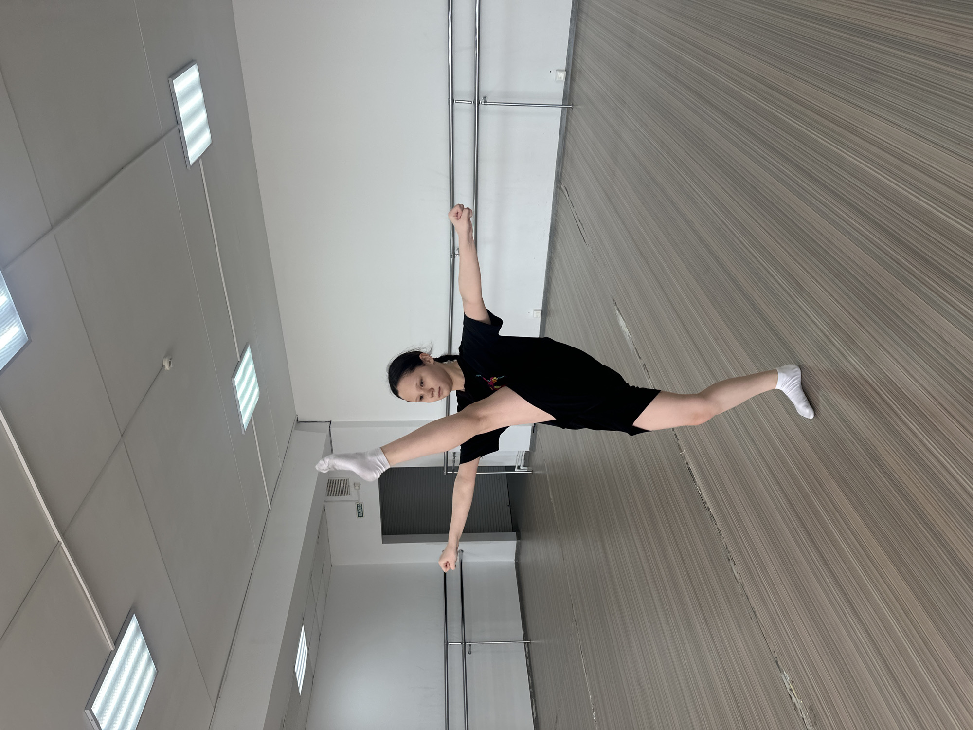 A young woman wearing a black T-shirt and shorts is doing a split in a dance studio. She is wearing white socks and has her arms raised in fists. The studio has a light grey floor with a wood grain pattern and white walls with fluorescent lights. There are ballet bars on the walls. The image was taken in Votkinsk, Russia.