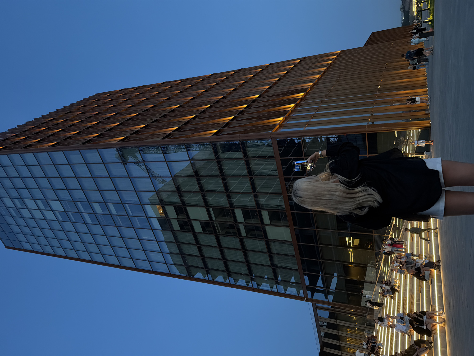 The image shows a tall, modern building with a glass facade and a metal roof structure, possibly the Kazan Family Center in Kazan, Russia. The building has a sharp, triangular shape, and the glass reflects the surrounding environment. The metal roof structure appears to have a pattern of illuminated openings. In the foreground, there are people standing on steps or a walkway, some of whom are seated. One woman with long blonde hair is facing the building and taking a picture with her phone. The sky is a deep blue, suggesting it is dusk or early evening.