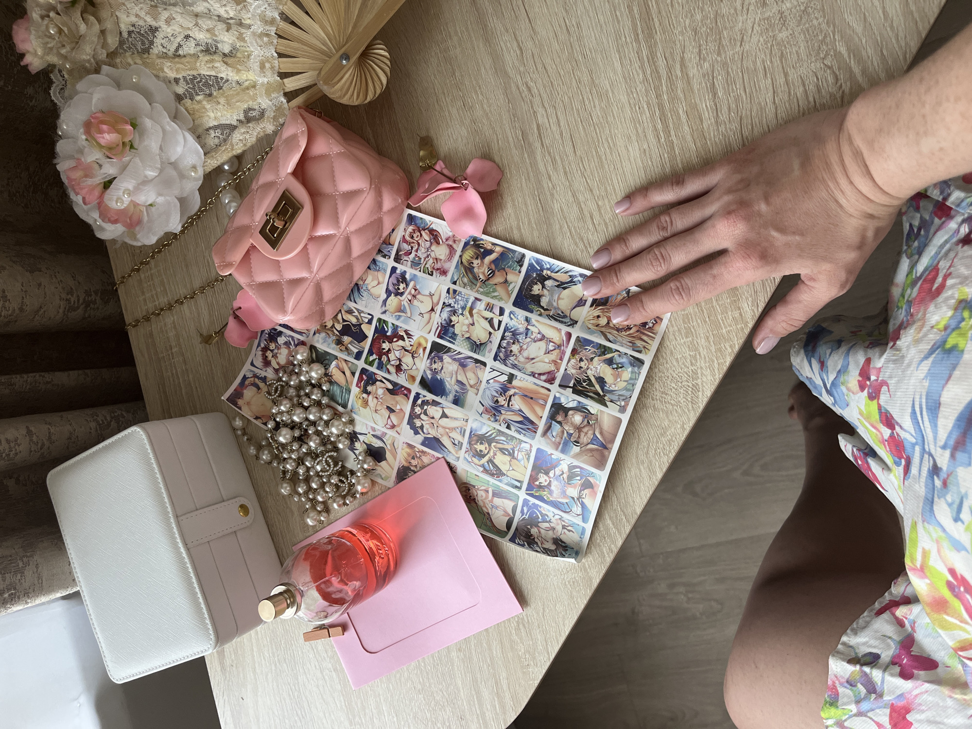 The image shows a flat lay of various feminine accessories and anime-style stickers arranged on a wooden surface. In the upper left is a white jewelry box. To its right is a pink envelope and a bottle of pink liquid, likely perfume. In the center of the image are a string of pearls and a sheet of stickers featuring anime characters in bikinis. To the right of the stickers is a small pink quilted handbag with a gold clasp. Further to the right is a decorative fan and a cluster of artificial flowers with pearl accents. A person's hand with light pink nail polish rests on the lower right side of the image, with their floral-patterned clothing visible in the bottom left corner. The scene is indoors, with a neutral-colored curtain visible in the background. There is no discernible indication of the specific location in Kazan, Russia.