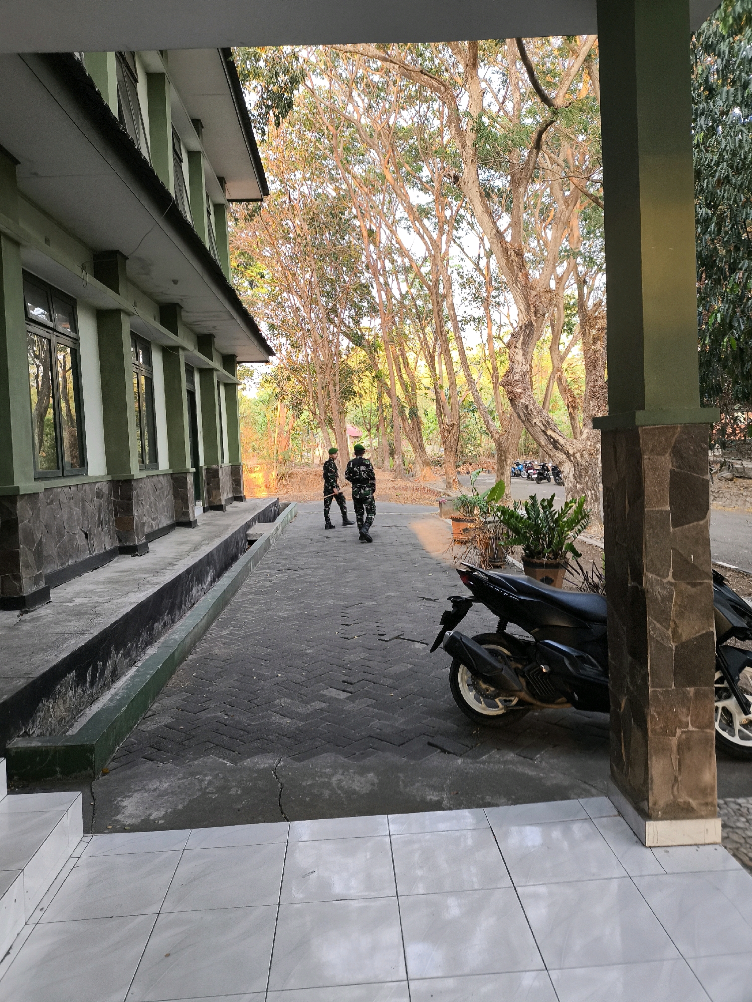 The image shows an outdoor scene in front of a military-style building. The building is light green with dark green accents around the windows and columns. The lower part of the building features a stone facade. In front of the building is a paved walkway made of interlocking bricks, with a drainage channel running alongside it. Two individuals in military uniforms are walking away from the camera on the walkway, one holding a long object that could be a rifle or a staff. A black motorcycle is parked on the right side of the walkway, partially obscuring a stone-clad pillar that supports an overhang. Beyond the walkway, there is a dense arrangement of trees with bare branches, suggesting a dry season or a specific type of deciduous tree. The lighting indicates it is either early morning or late afternoon, with warm sunlight filtering through the trees. In the background, several other motorcycles are parked, and the environment appears somewhat unkempt, with dry leaves on the ground. The scene is consistent with a military compound or training area.