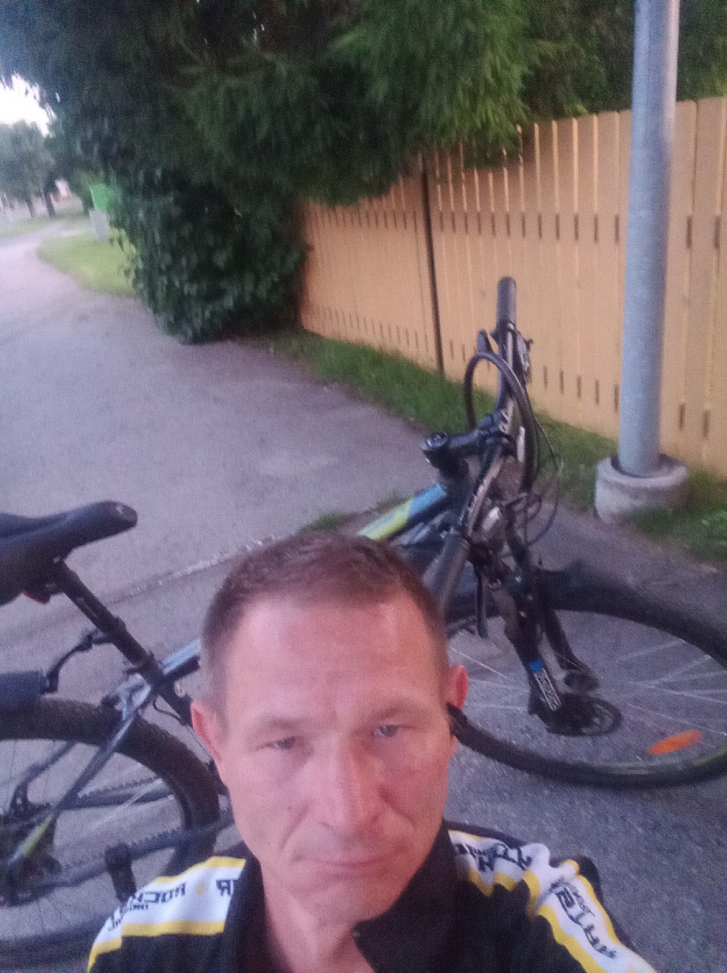 A man's face is prominently featured in the foreground of this outdoor image, with the lower portion of his body and a bicycle behind him. The man has short, light brown hair and is looking directly at the camera with a neutral expression. He is wearing a black cycling jacket with yellow and white detailing and what appears to be an earbud in his right ear.
To the left and behind the man is the rear of a bicycle, showing its saddle, seat post, and part of the rear wheel. The bicycle frame is visible, with blue and yellow accents. The front of the bicycle is also partially visible, including the handlebars, brake levers, and front wheel. The bicycle appears to be a mountain bike.
The environment suggests an urban or suburban setting in Tapa, Estonia. A paved pathway runs from the foreground into the background, curving to the left. To the right of the pathway is a light yellow wooden fence with vertical planks. Behind the fence, lush green trees and bushes form a dense backdrop. A grey cylindrical lamppost is situated next to the fence in the mid-ground. The ground is a mix of pavement and grass. The lighting suggests it is late afternoon or early evening.