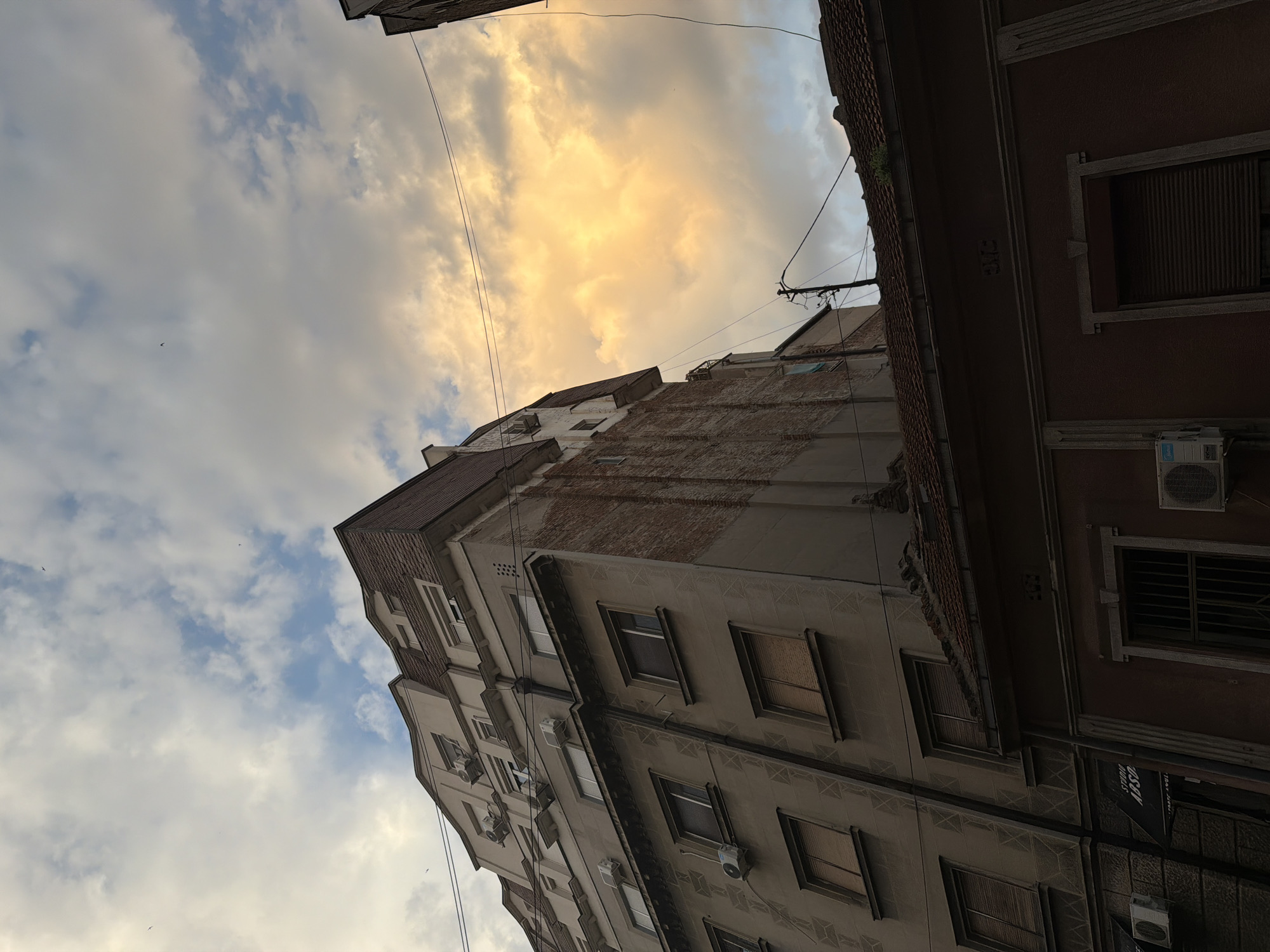 The image depicts a view of buildings and sky in Vracar Urban Municipality, Serbia. The architecture is characterized by multi-story buildings with visible brickwork and various windows, some with closed blinds. Air conditioning units are mounted on the exterior walls of some structures. Power lines and a utility pole are present, crisscrossing the sky. The sky is filled with clouds, with a prominent area of bright, yellowish-orange clouds suggesting sunset or sunrise lighting. Several small birds are visible in the sky. The lower portion of the image shows the roofline of a lower building, with red tiles and some greenery growing on the edge. The scene appears to be an urban setting captured from a street-level perspective, looking upwards towards the sky and the upper levels of the buildings.