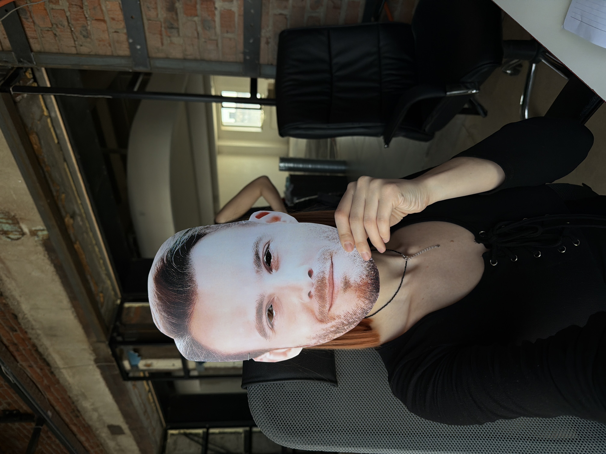 The photograph captures an individual holding a printed face mask in front of their own face. The person is wearing a black, long-sleeved shirt and a thin necklace with a pendant. The setting seems to be an interior space with exposed brick walls and metal beams, possibly an office or studio. Behind the person, there's an office chair and a desk with papers on it. The photograph is taken at an angle, making the room appear rotated. There are no visible elements that would indicate the location as the city of Chaikovsky, Russia.