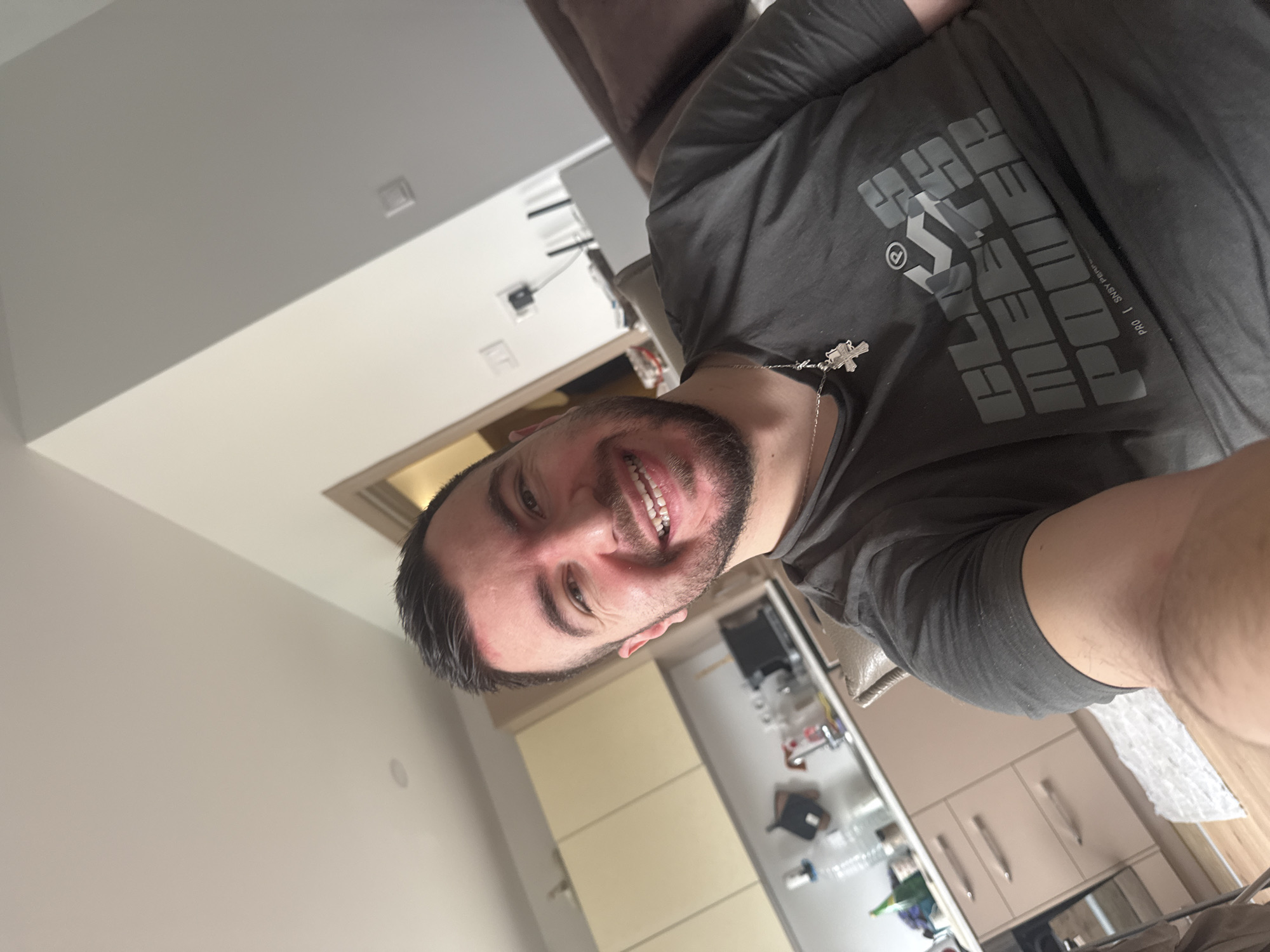 A person is seen in an angled indoor shot. The person has dark hair, a beard, and is wearing a grey t-shirt with lettering and a silver cross necklace. The background includes a kitchen area with light-colored cabinets, a countertop with various items, and a white wall with light fixtures. A beige sofa is partially visible at the top of the frame. The location is likely Sofia, Bulgaria.