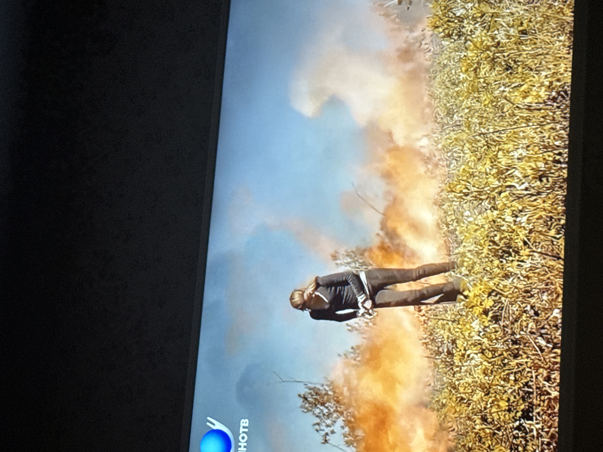 The image shows a scene from a television screen. The scene depicts a woman standing in a field with orange smoke. The background includes a blue sky, shrubbery, and grass. A television logo is visible at the bottom left of the screen. There is no evidence to suggest the location is Mytishchi, Russia.