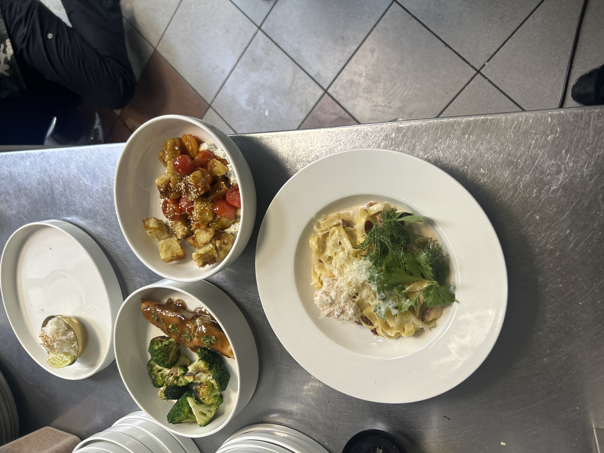 The image shows a culinary setup with three bowls of food and a plate of pasta, arranged on a stainless steel counter. In the top bowl, there are bite-sized pieces of fried food, possibly tofu or chicken, mixed with cherry tomatoes in a glossy sauce, sprinkled with sesame seeds. To the left and slightly below, another bowl contains a piece of glazed salmon served with steamed broccoli florets. Further left, on a large white plate, is a small mound of white rice in a golden dish, accompanied by a lime wedge. The largest dish, on the right, is a white plate filled with fettuccine pasta in a creamy sauce, topped with shredded cheese and fresh herbs like dill and parsley. The background reveals a tiled floor with a geometric pattern and a glimpse of a person wearing black pants, suggesting a kitchen or restaurant environment. The lighting appears to be artificial, consistent with an indoor setting. The scene depicts food preparation or presentation, with a focus on the dishes themselves. No visible text is present in the image.