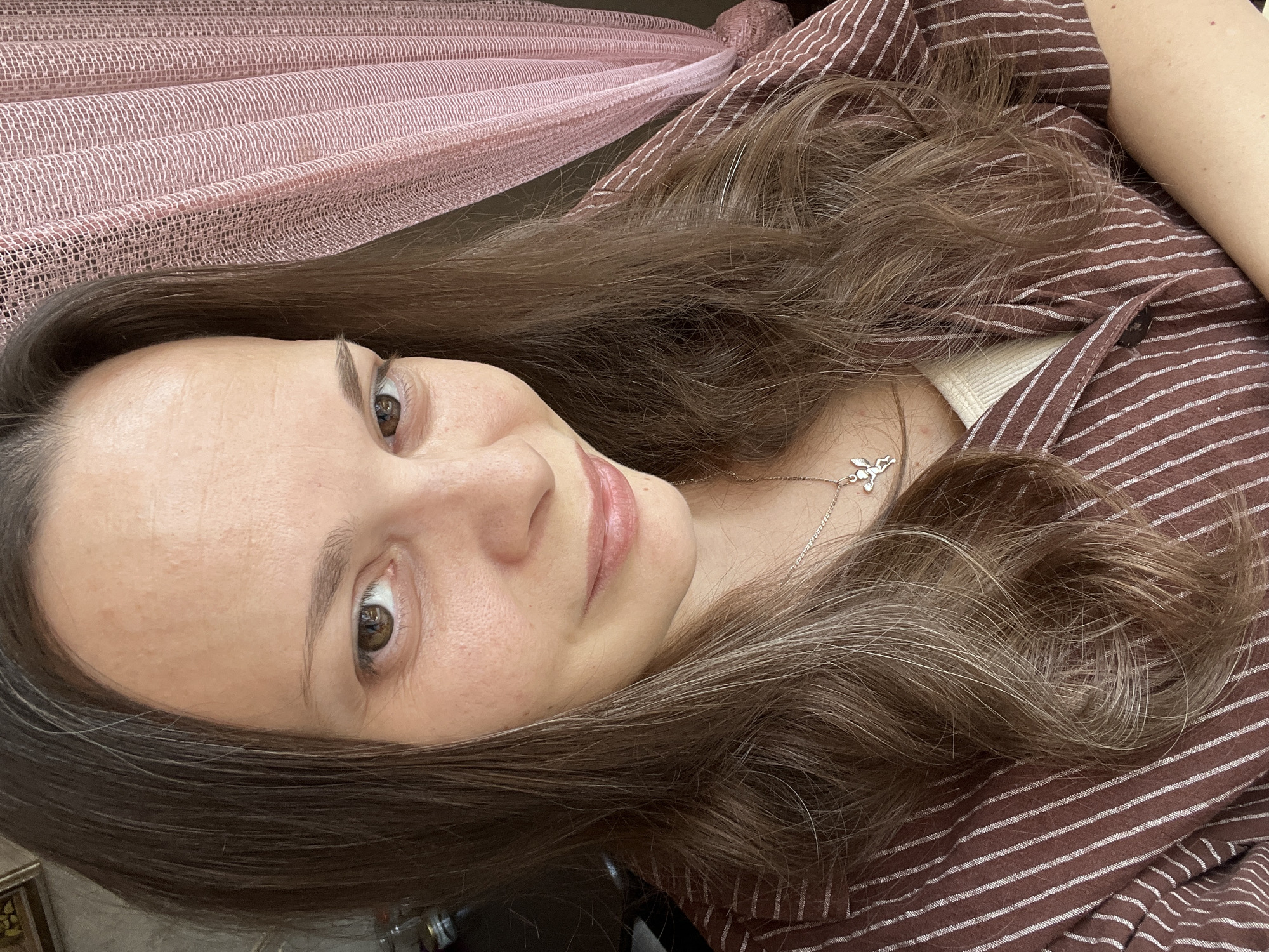 A woman with long, wavy brown hair and brown eyes is captured in a close-up, selfie-style photograph. She is wearing a brown and white striped shirt over a cream-colored top, and a silver pendant with a winged figure is visible around her neck. She has a subtle smile and her head is tilted slightly. To her right, a textured pink curtain is visible. The background appears to be an indoor setting. No specific location in Tolyatti, Russia can be identified from the visual information provided.