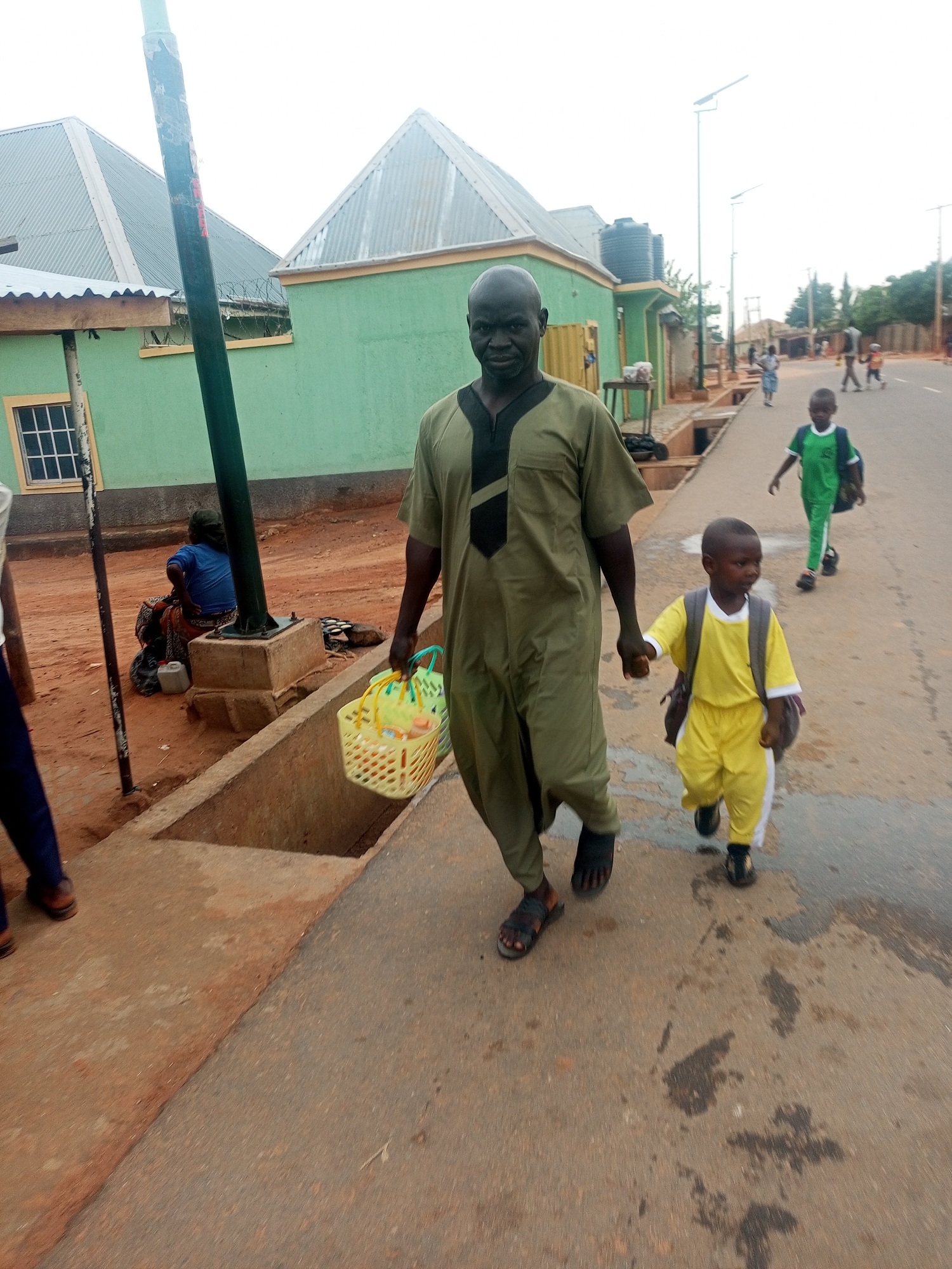 An adult male and a young boy are walking hand-in-hand on a paved surface in the city of Gombe, Nigeria. The adult is wearing an olive green robe with a black trim at the neck and sandals. He is carrying a yellow plastic basket filled with assorted items. The boy is wearing a yellow tracksuit with a white collar and white stripes down the sides. He is also wearing black shoes and carrying a backpack.
In the background, there is a green building with a corrugated metal roof and a square window. A woman is seated on the ground near the building. Further down the street, another child is walking in the same direction. The road is paved and there are a few other people visible in the distance. Modern streetlights are visible along the side of the road.