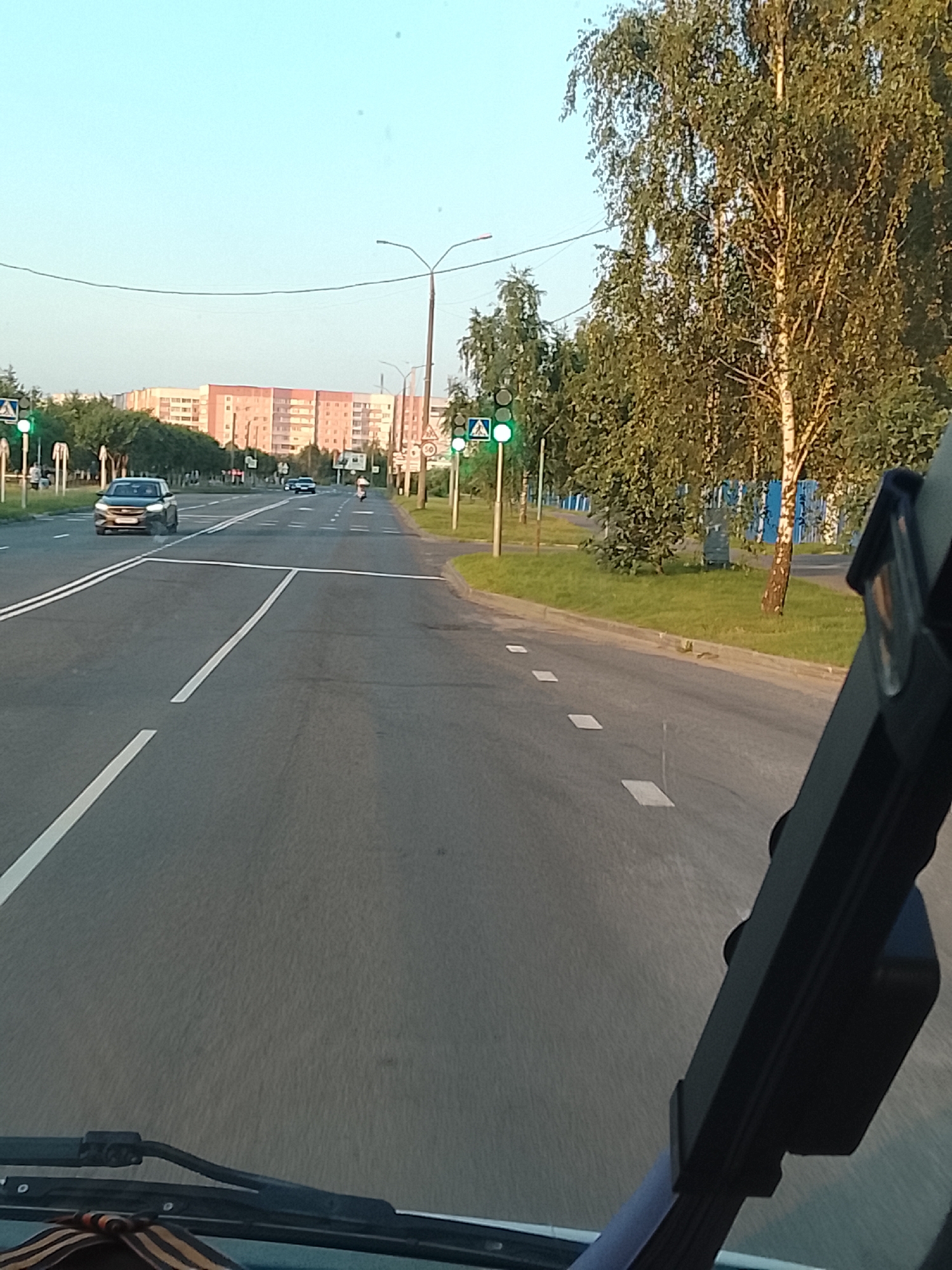 The image shows a view from inside a vehicle, likely a car, looking out at a street in Zhlobin, Belarus. The road is paved and has clear lane markings. There are several cars on the road, including a dark-colored sedan in the left lane and other vehicles further ahead. Traffic lights are visible, with one showing a green light. Pedestrian crossings are marked on the road with white squares. Trees line the right side of the road, and a tall, slender birch tree is prominently featured. In the background, there are multi-story residential buildings with a pinkish-orange facade. The sky is clear and blue, indicating daylight. The scene appears to be captured during the daytime, possibly late afternoon, given the lighting conditions.