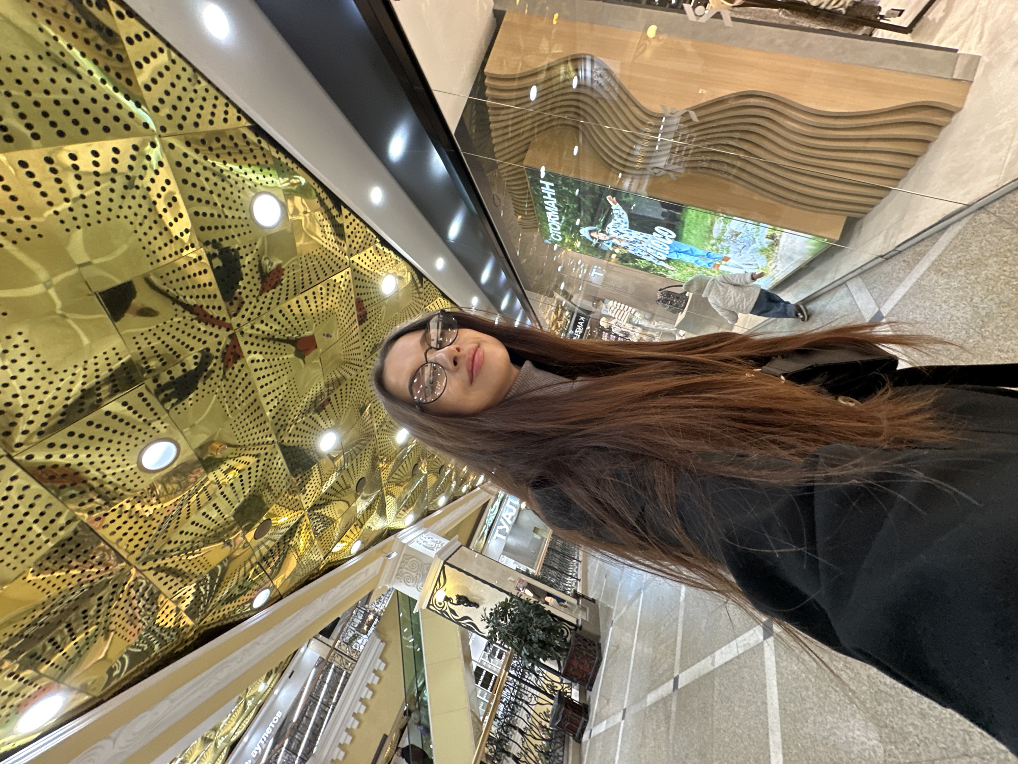 The image is a dynamic, diagonally-oriented selfie taken by a young woman with long, straight brown hair and clear-framed glasses, looking directly at the camera with a slight smile. She is wearing a grey turtleneck or sweater underneath a black jacket or coat.
The setting is an opulent, brightly lit indoor shopping mall or complex, confirmed to be in Yekaterinburg, Russia. A striking feature of the interior is the ceiling, which is covered in numerous golden, reflective, and perforated panels that create a complex, geometric, and somewhat futuristic pattern. Several circular recessed lights are visible on this ceiling.
In the background, behind a clear glass barrier, there are multi-level architectural structures with cream-colored ornate railings and balustrades, suggesting balconies or upper floors. One prominent shop front features a large display window with a distinctive wooden, wave-like paneling and a large screen showing a person in outdoor attire. Text visible on this shop display includes "СТОКМАНН" (Stockmann), a well-known department store, and possibly "ОNINENН". Another fragmented piece of Cyrillic text, "...ть ауметов", can be seen on a white column or sign in the lower right. A small potted plant and an intricate metal railing are also visible near what appears to be an escalator or staircase.
The overall activity is the woman taking a selfie, capturing herself within this unique and grand shopping environment. The artificial lighting indicates an active indoor space, likely during the day. The scene combines modern design with classical architectural accents, reflecting a high-end commercial establishment.